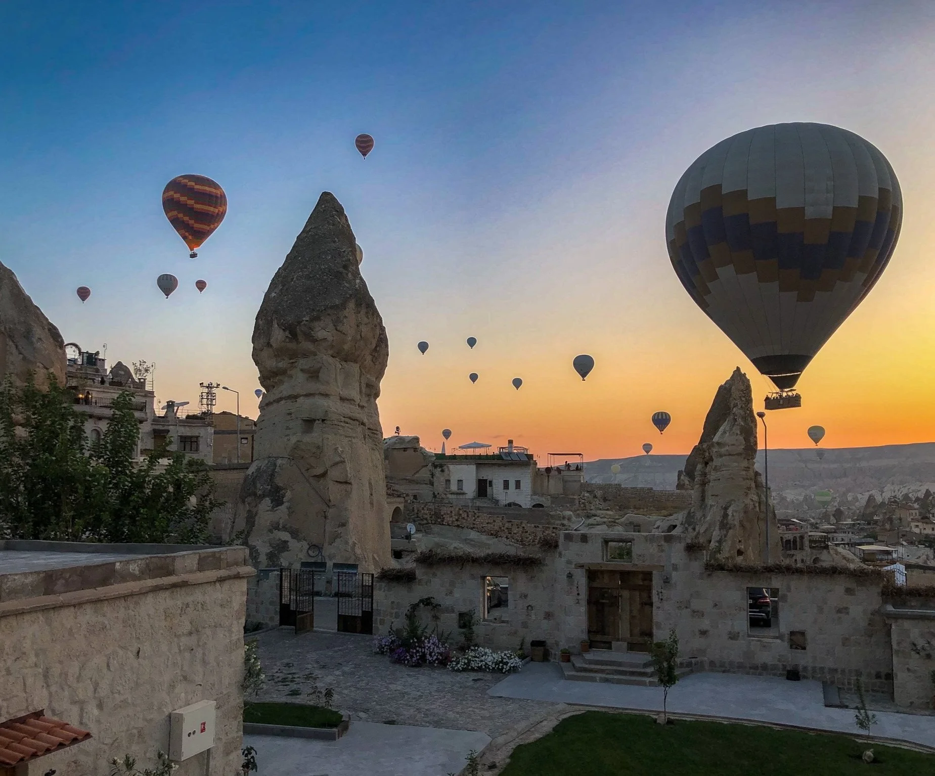 Multiple hot air balloons floating over rock formations and historic stone buildings during a sunset in Cappadocia, Turkey.