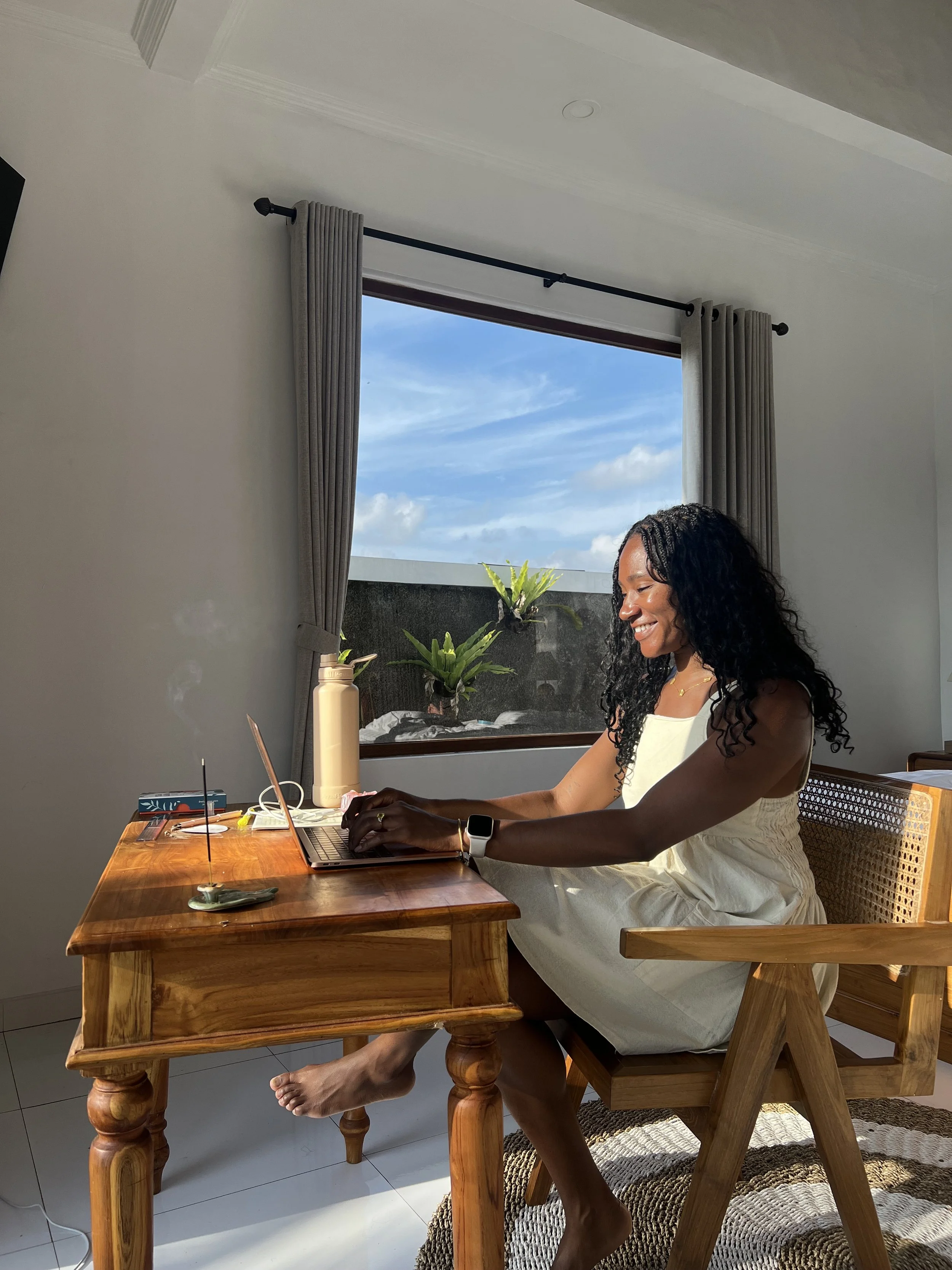 A Black woman sitting at a wooden desk working on a laptop, with sunlight coming through a window behind her, during daytime.