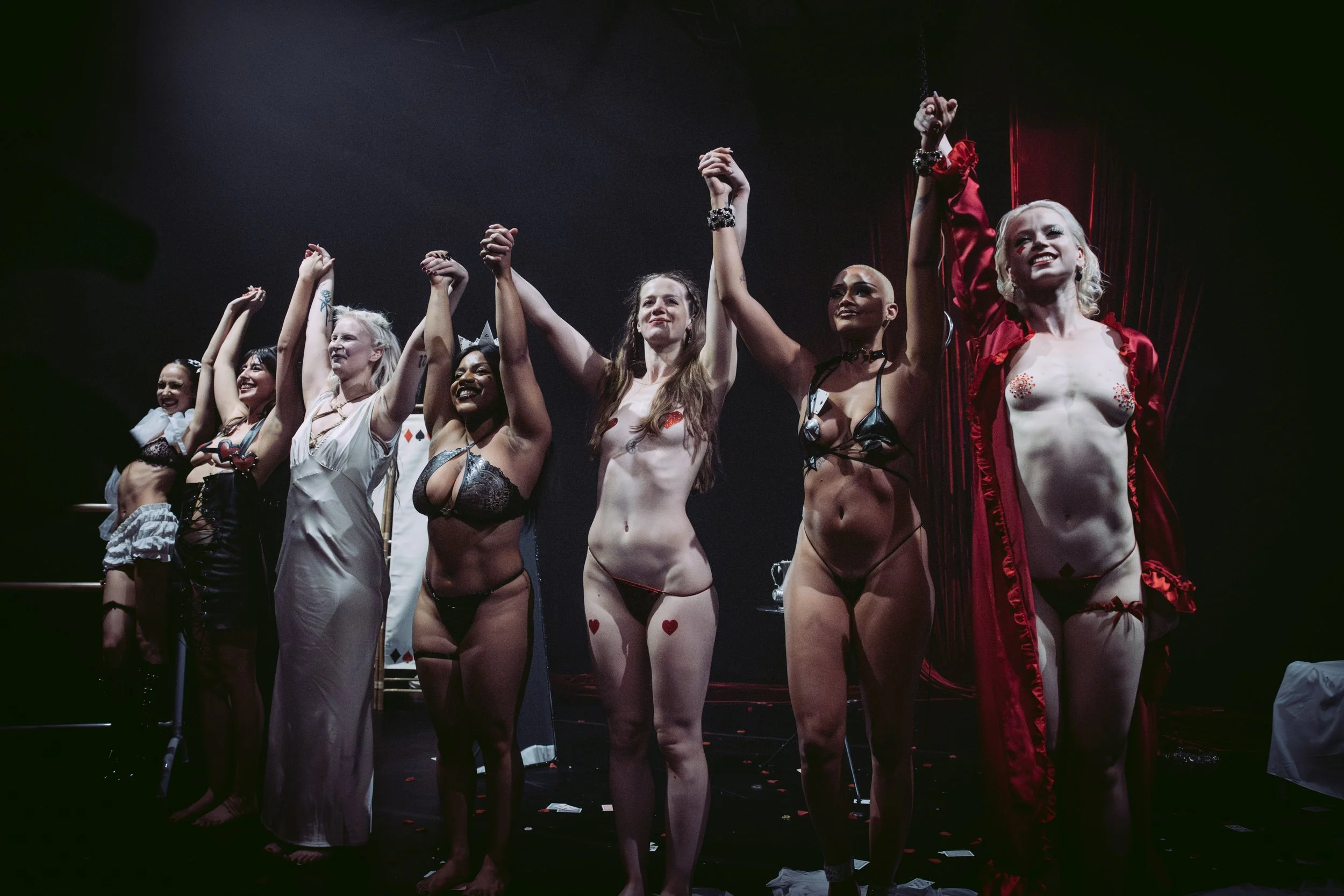 Group of women with diverse skin tones dressed in lingerie or costumes, standing on stage with arms raised and holding hands, smiling and celebrating.