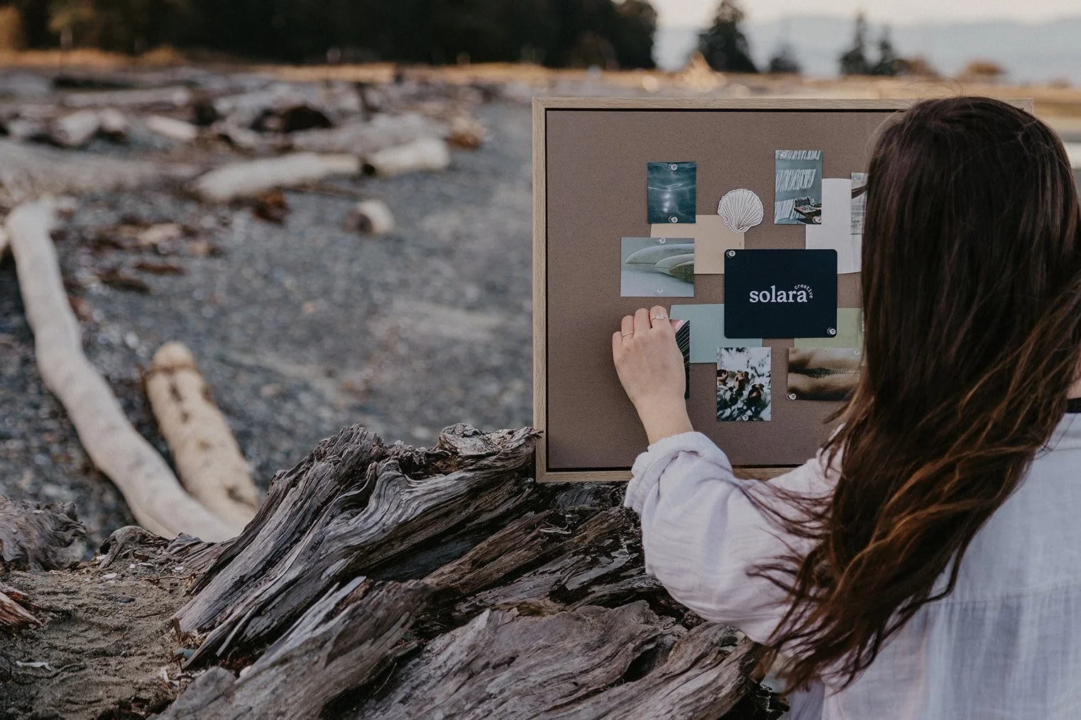 A person pins inspiration images featuring blue and green tones and water reflections to a vision board on the beach during a Solara Creative strategy session, signifying a clean, meaningful brand.