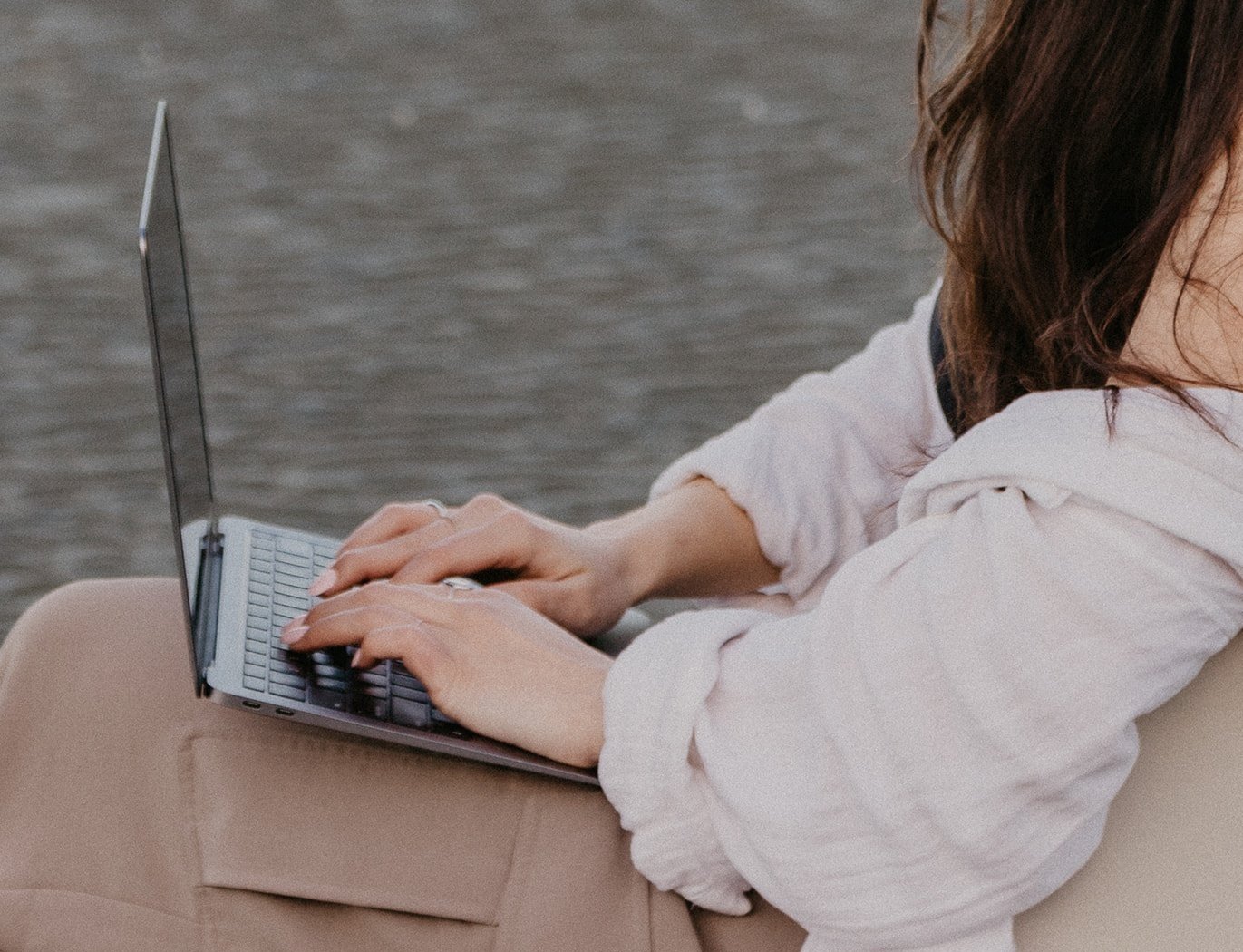 A grainy aesthetic photo of a person in a loose white shirt, black tank top, and beige pants typing on a laptop while writing their brand story on a sandy beach.