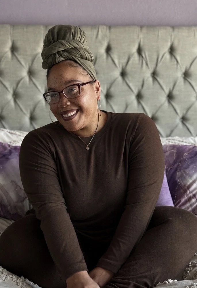 A woman with glasses, a head wrap, and dark lipstick sitting on a bed with a tufted headboard and purple pillows, smiling at the camera.