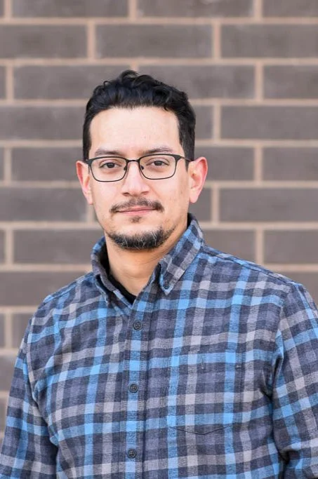 A man with glasses and a beard wearing a blue and gray plaid shirt standing outdoors in front of a brick wall.