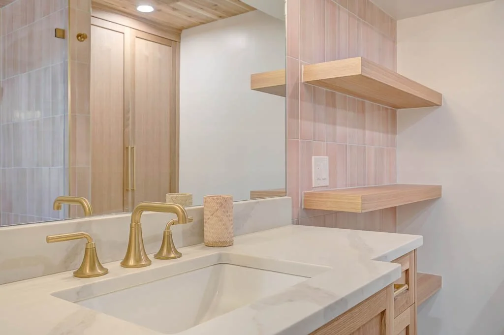 Bathroom sink with gold fixtures, a large mirror, and wooden shelves on a pink tiled wall