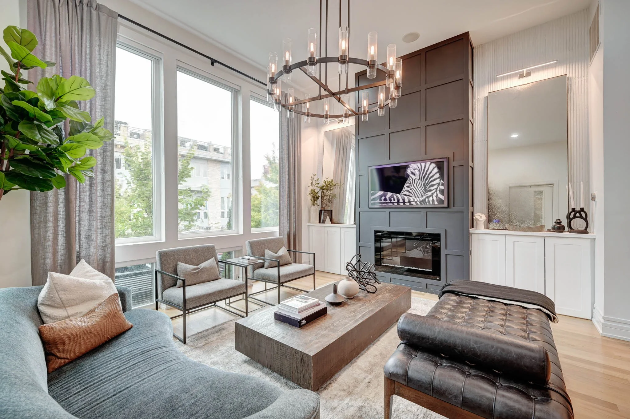Modern living room with large windows, gray sofa, beige chairs, a wooden coffee table, a dark leather bench, a fireplace, a mounted television, a chandelier, and decorative items.