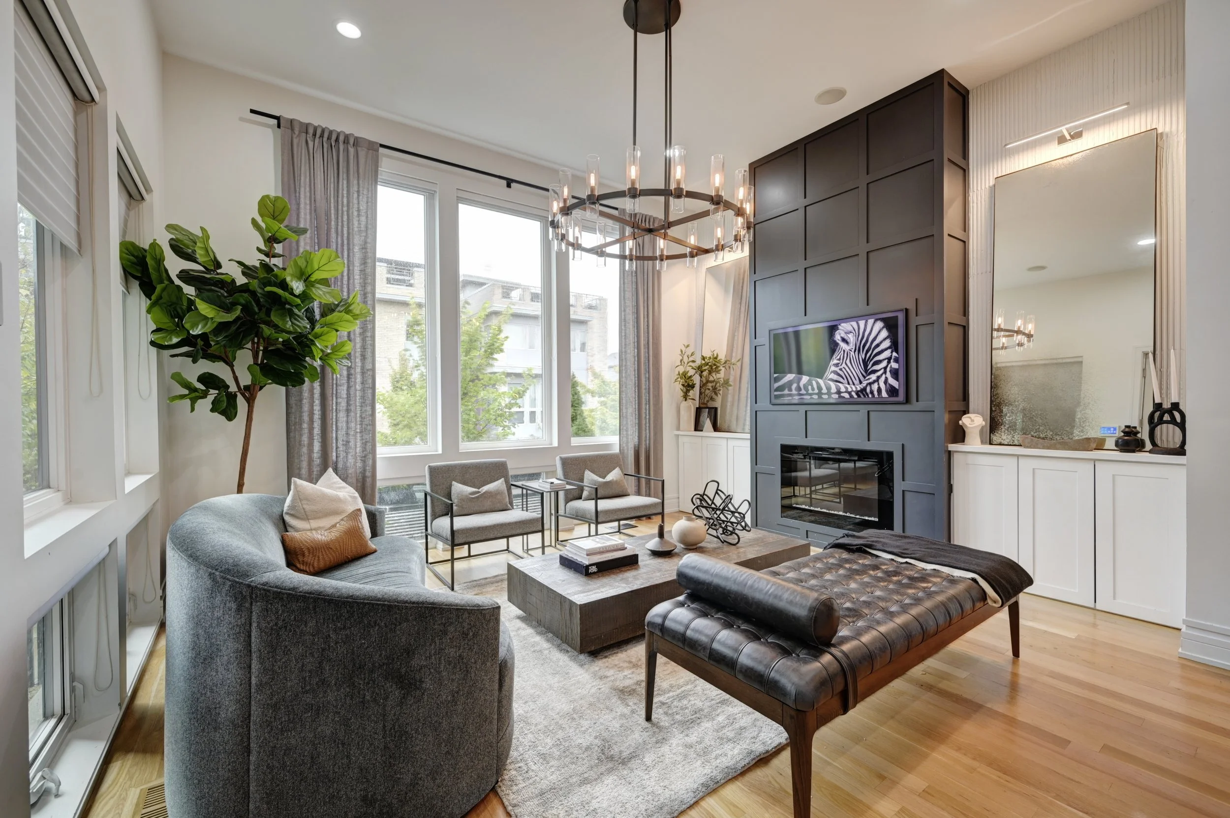Living room with large windows, a chandelier, a fireplace with a zebra print on the television screen, and various furniture including a gray sofa, armchairs, and a leather bench.