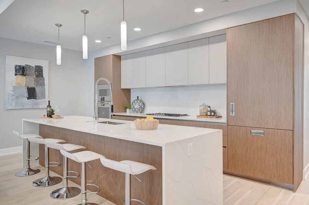 Modern kitchen with white and wood cabinetry, bar seating with four white stools, pendant lights, and abstract wall art.