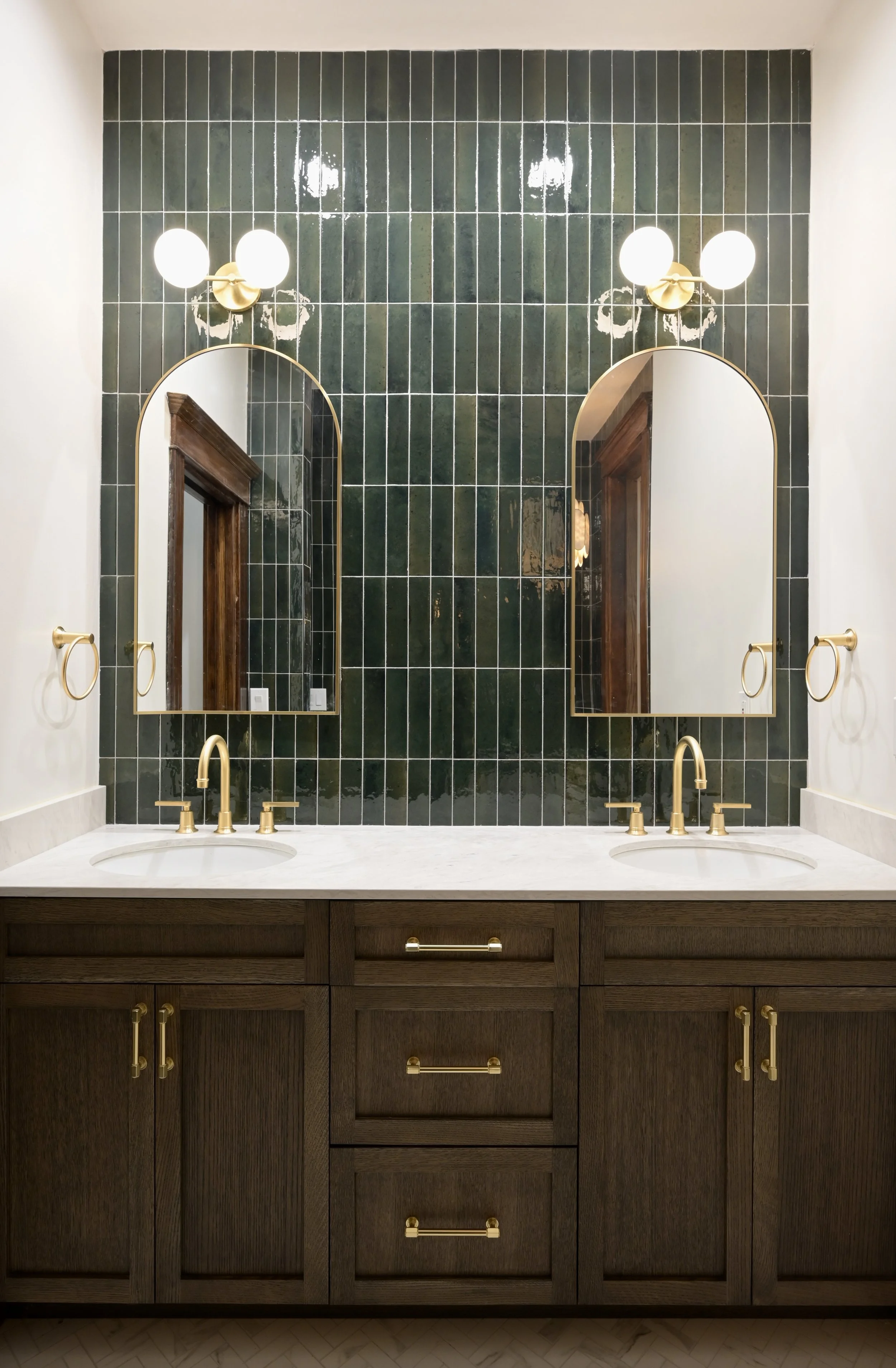 A bathroom vanity with two sinks, dark wood cabinets, gold fixtures, and two matching mirrors above each sink. The wall behind is tiled dark green, accented with wall-mounted light fixtures.
