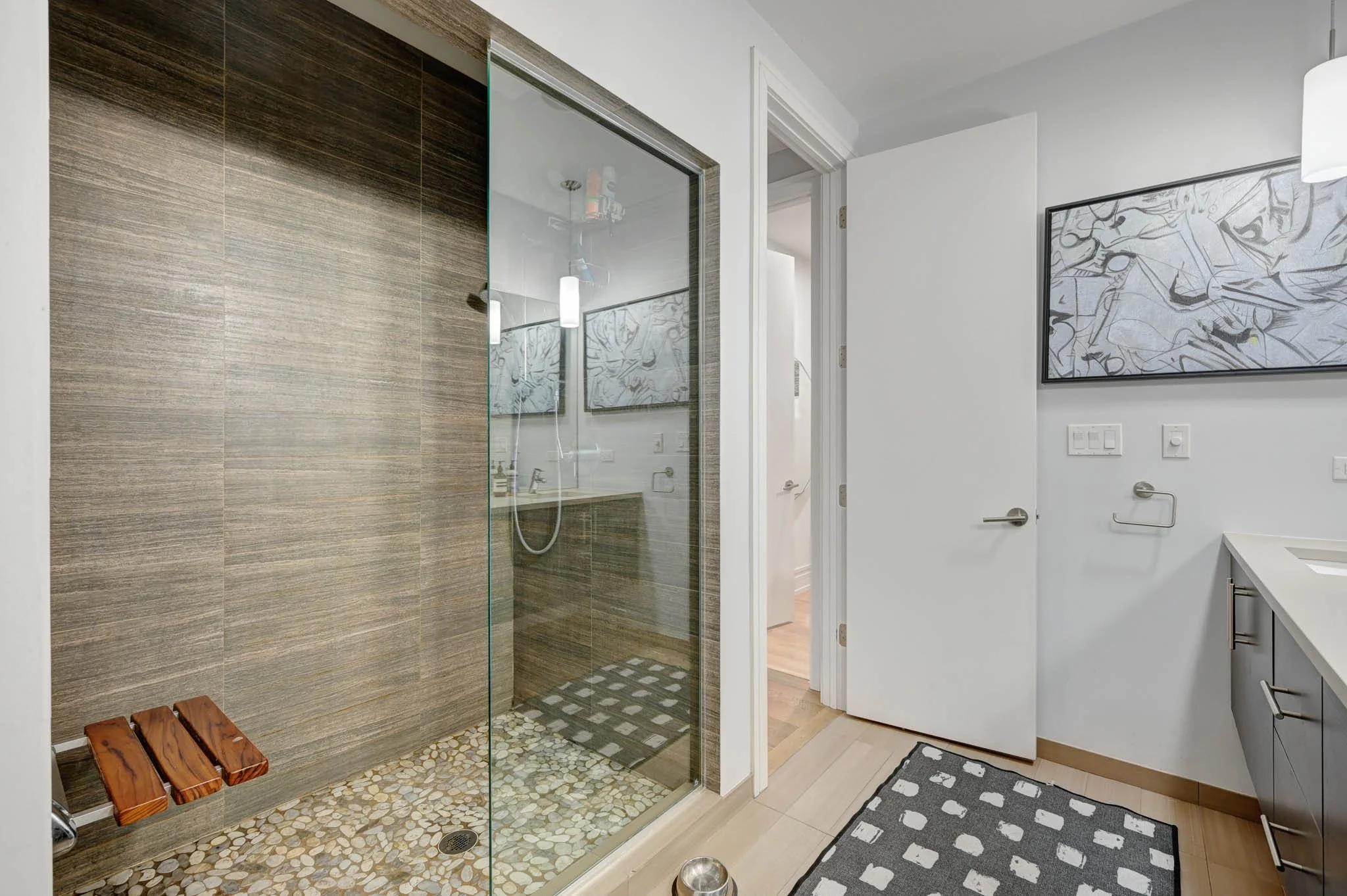 A modern bathroom with a glass shower enclosure, wooden bench, black and white abstract art on the wall, and a gray cabinet with a patterned rug.