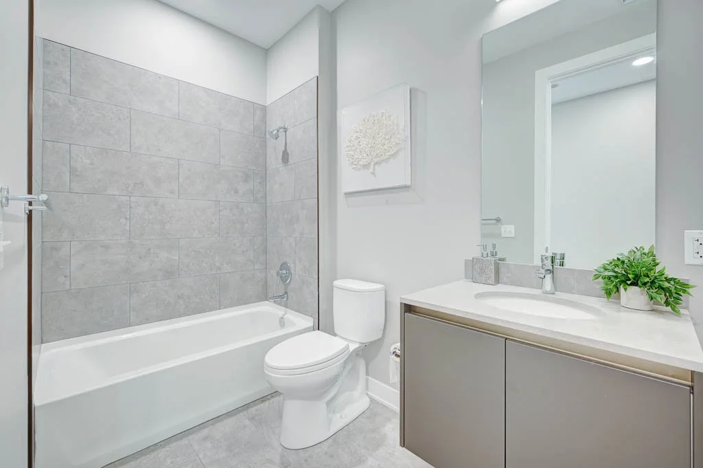 Modern bathroom with gray tiled walls, white bathtub, toilet, and vanity with a large mirror, small green plant on the sink, and a decorative wall art piece.