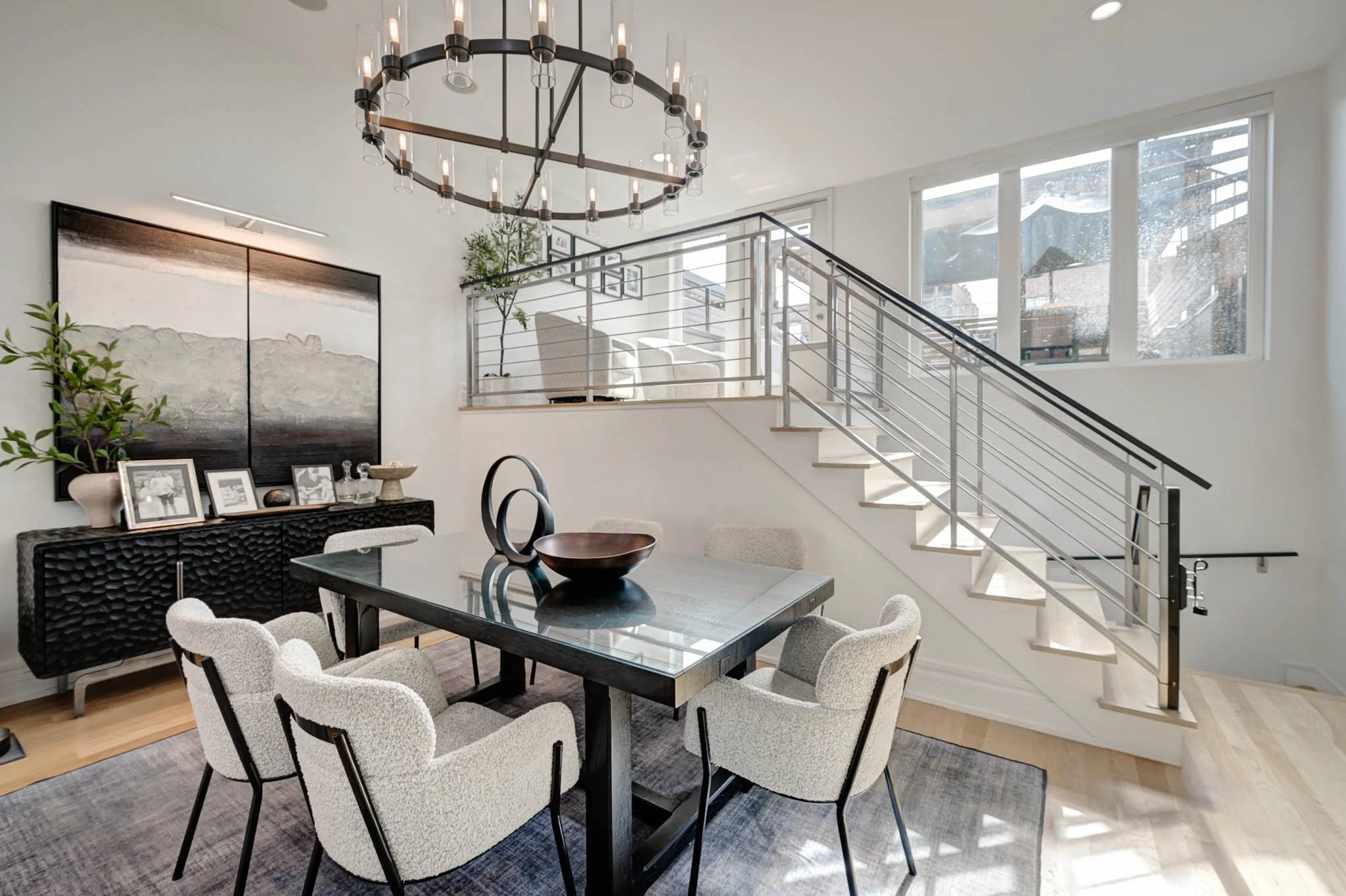 Modern dining area with a black rectangular table surrounded by cream-colored upholstered chairs, a large window bringing in natural light, a staircase with metal railing, and a chandelier with multiple exposed bulbs.