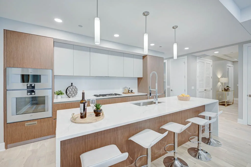 Modern kitchen with white countertops, wooden cabinets, and white upper cabinets. Four white bar stools at the island, pendant lighting, and built-in appliances.