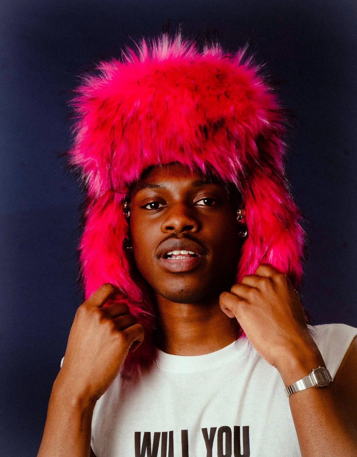 A person with dark skin wearing a bright pink furry hat that covers their head and ears, holding the sides of the hat with both hands, against a dark background.