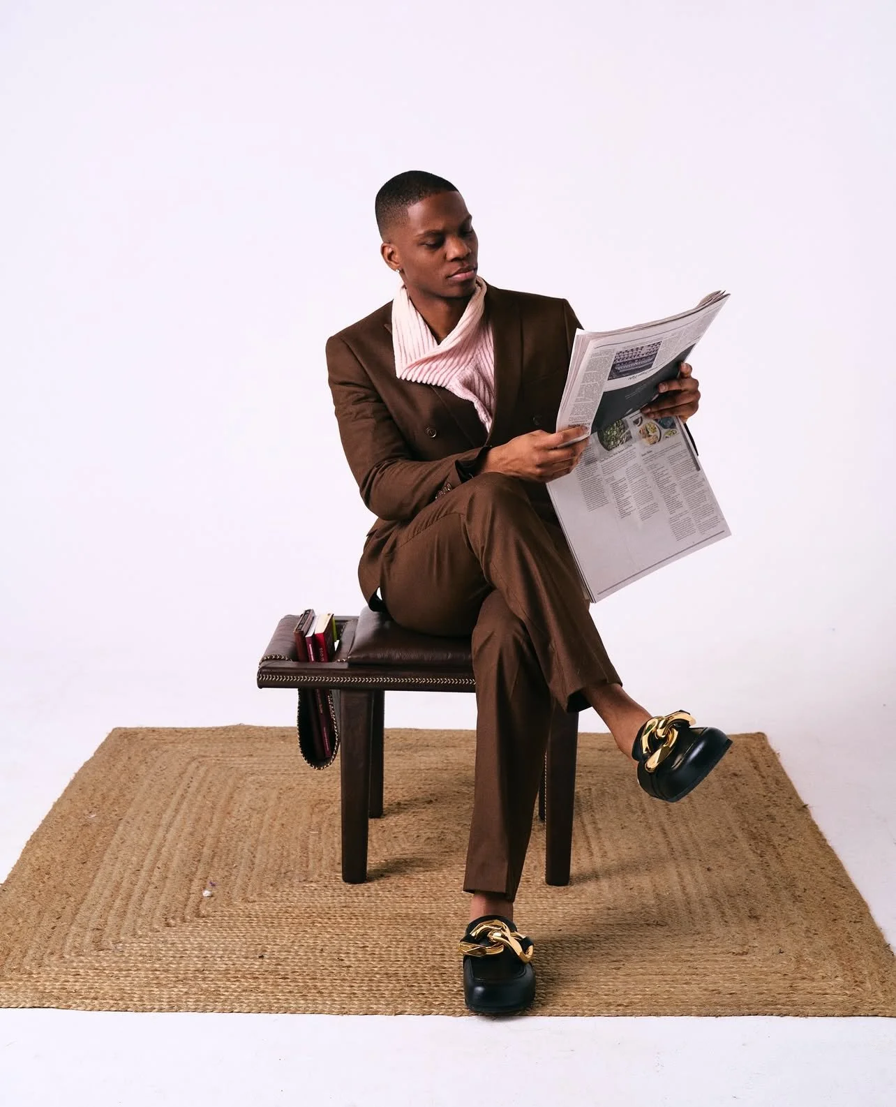 A woman in a brown suit with a pink shirt sits on a wooden bench, reading a newspaper, with a small stack of books beside her on the bench, on a beige rug against a plain white background.