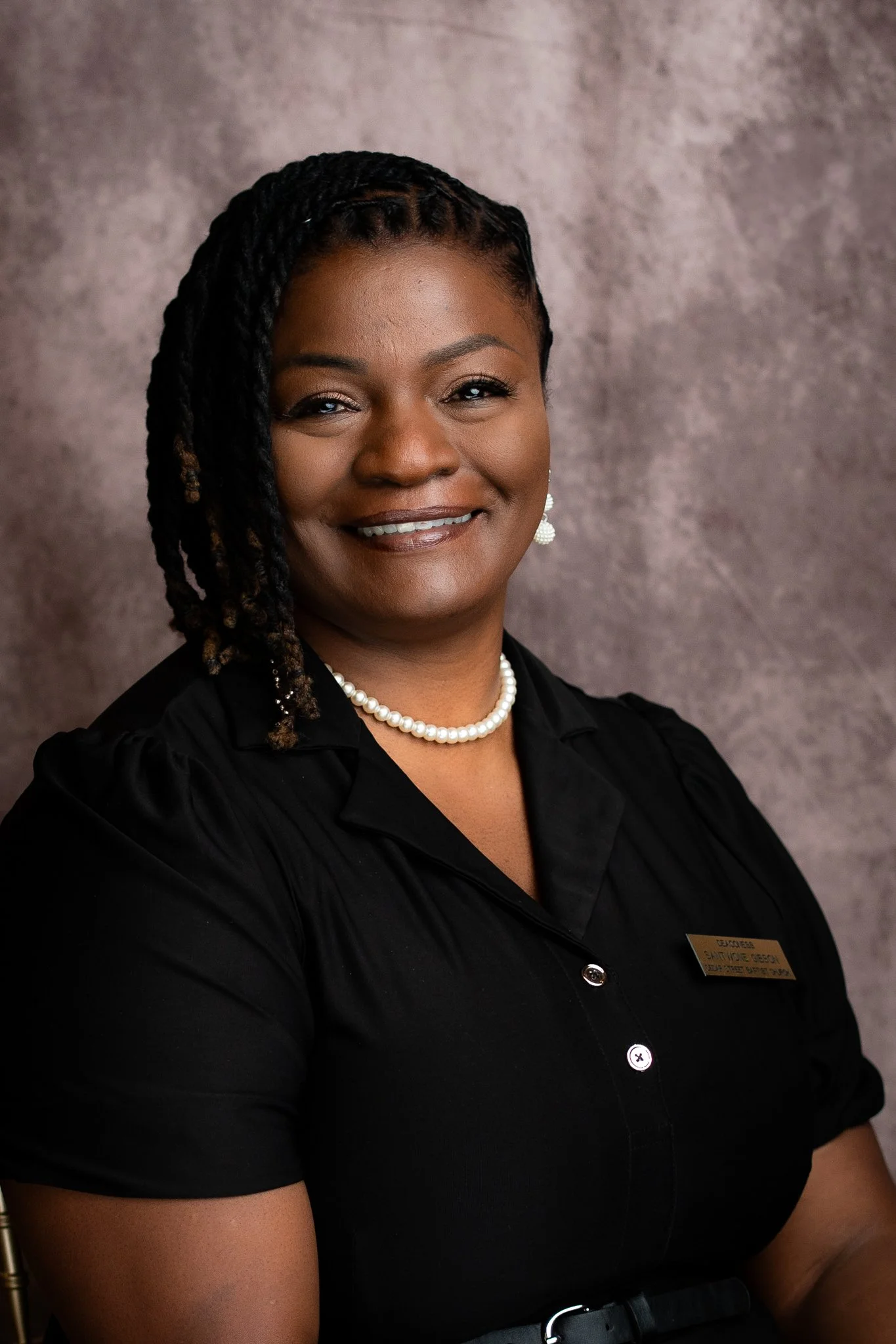 A professional woman with braided hair, wearing a black shirt, pearl necklace, and earrings, smiling in front of a textured background.