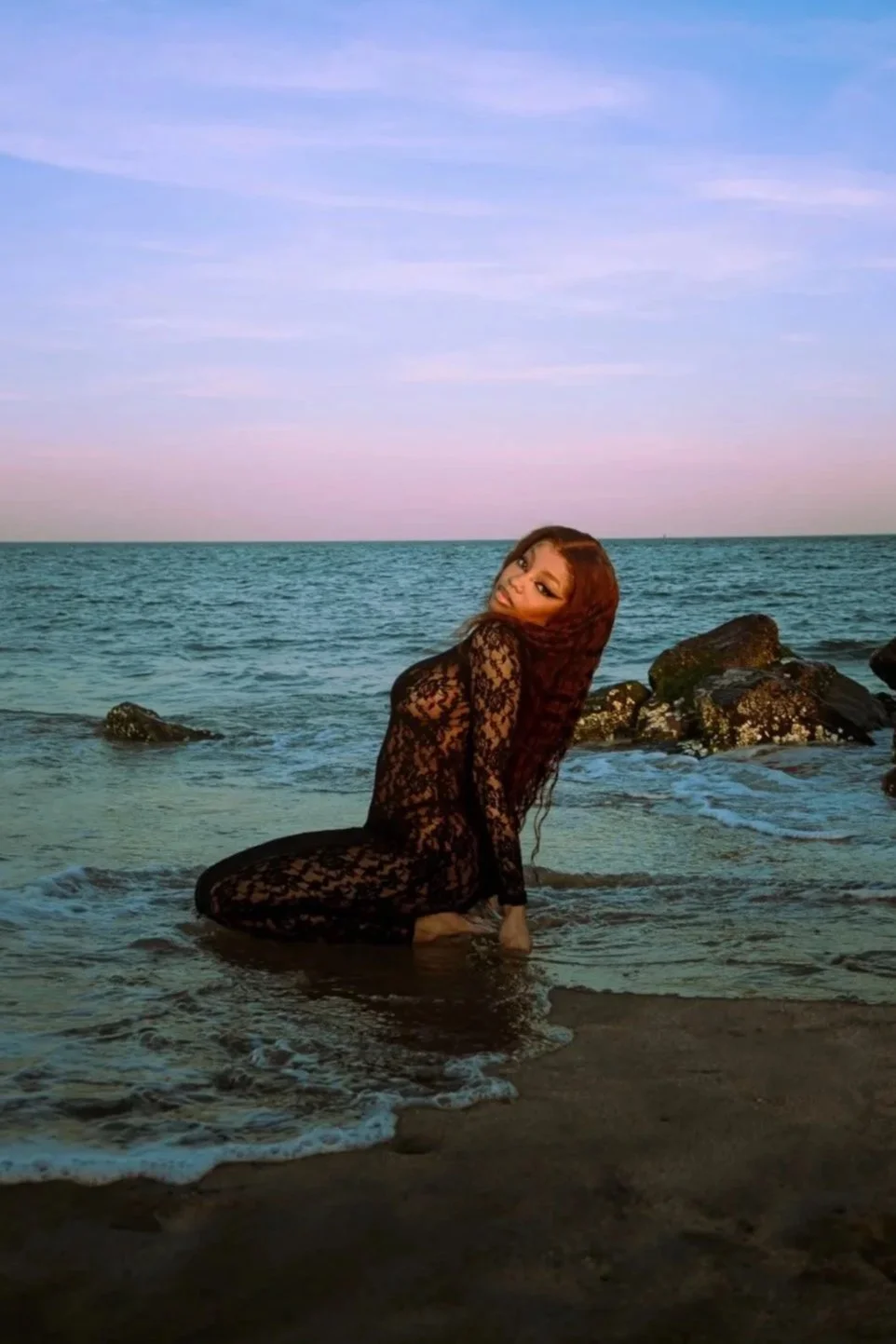 A woman with long, wavy hair wearing a black lace dress kneeling in shallow ocean water near rocks during sunset or sunrise.