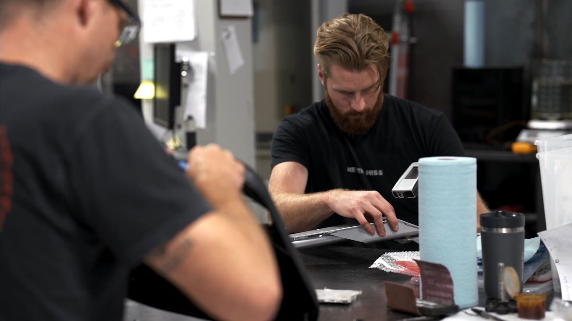 A man with a beard working on a laptop at a cluttered workbench in a workshop, with various tools and supplies around him.