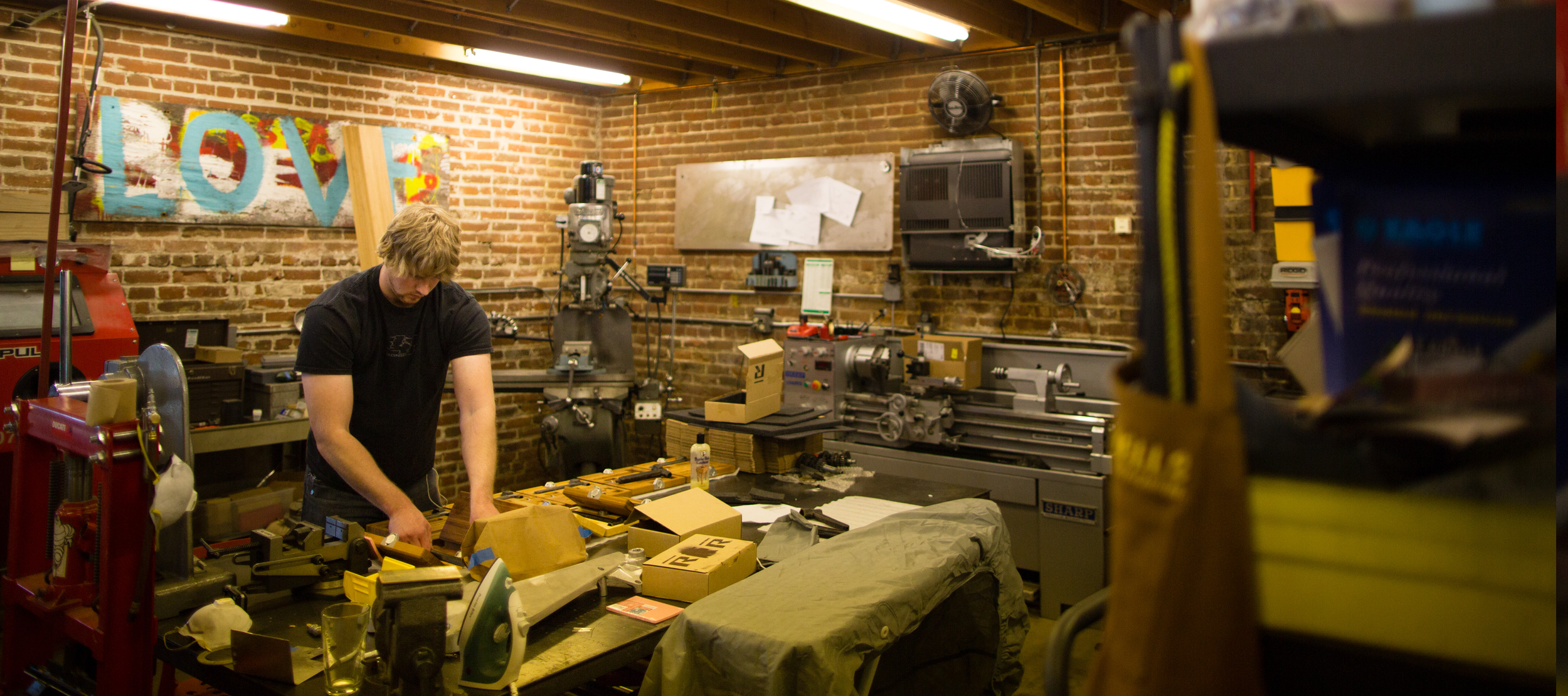A man working at a cluttered workbench in a workshop with exposed brick walls, surrounded by machinery, tools, and boxes.