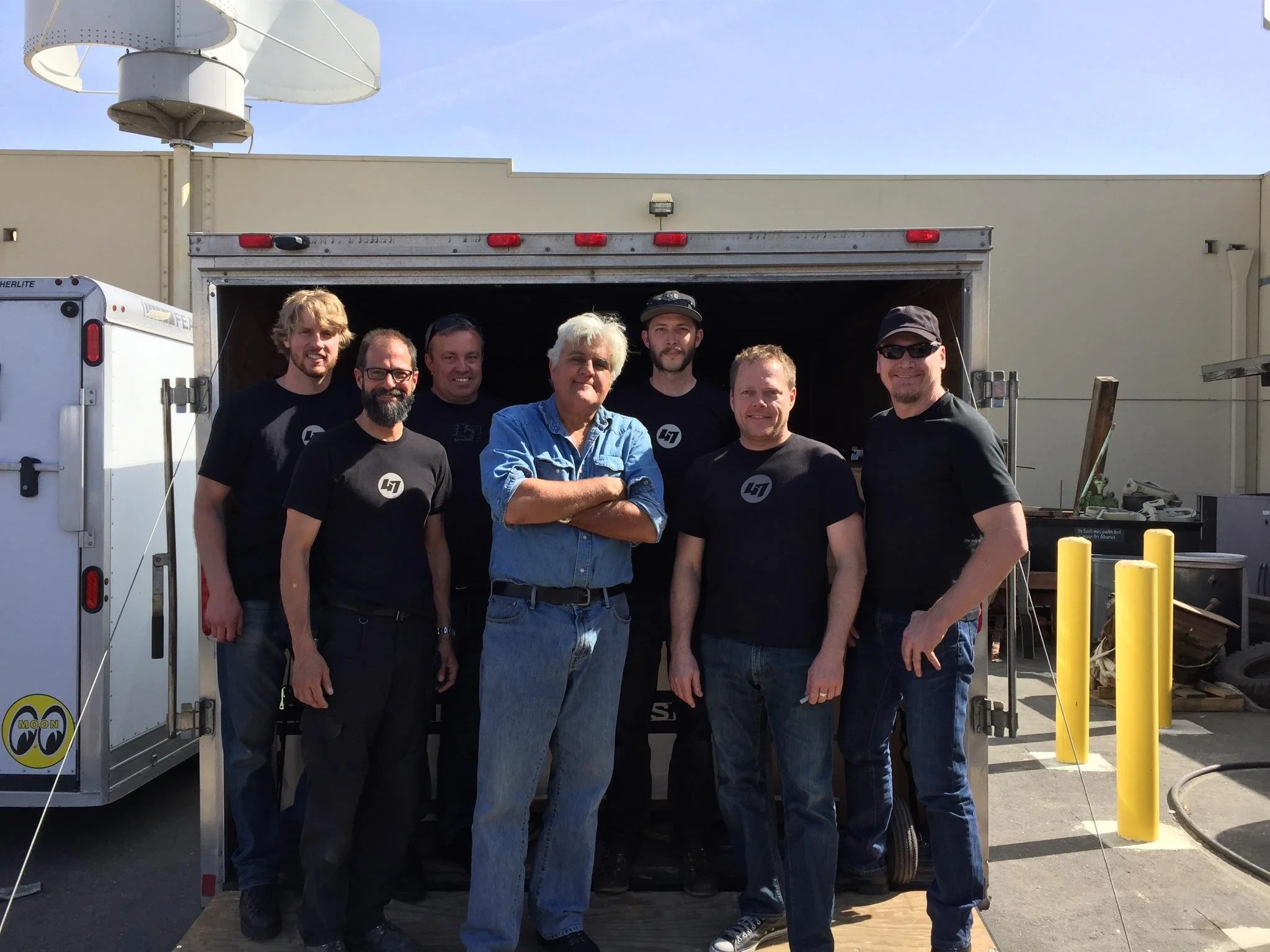 Group of eight men posing in front of a trailer, three are wearing black shirts with a logo, and one is in a blue denim shirt, outdoors on a sunny day.