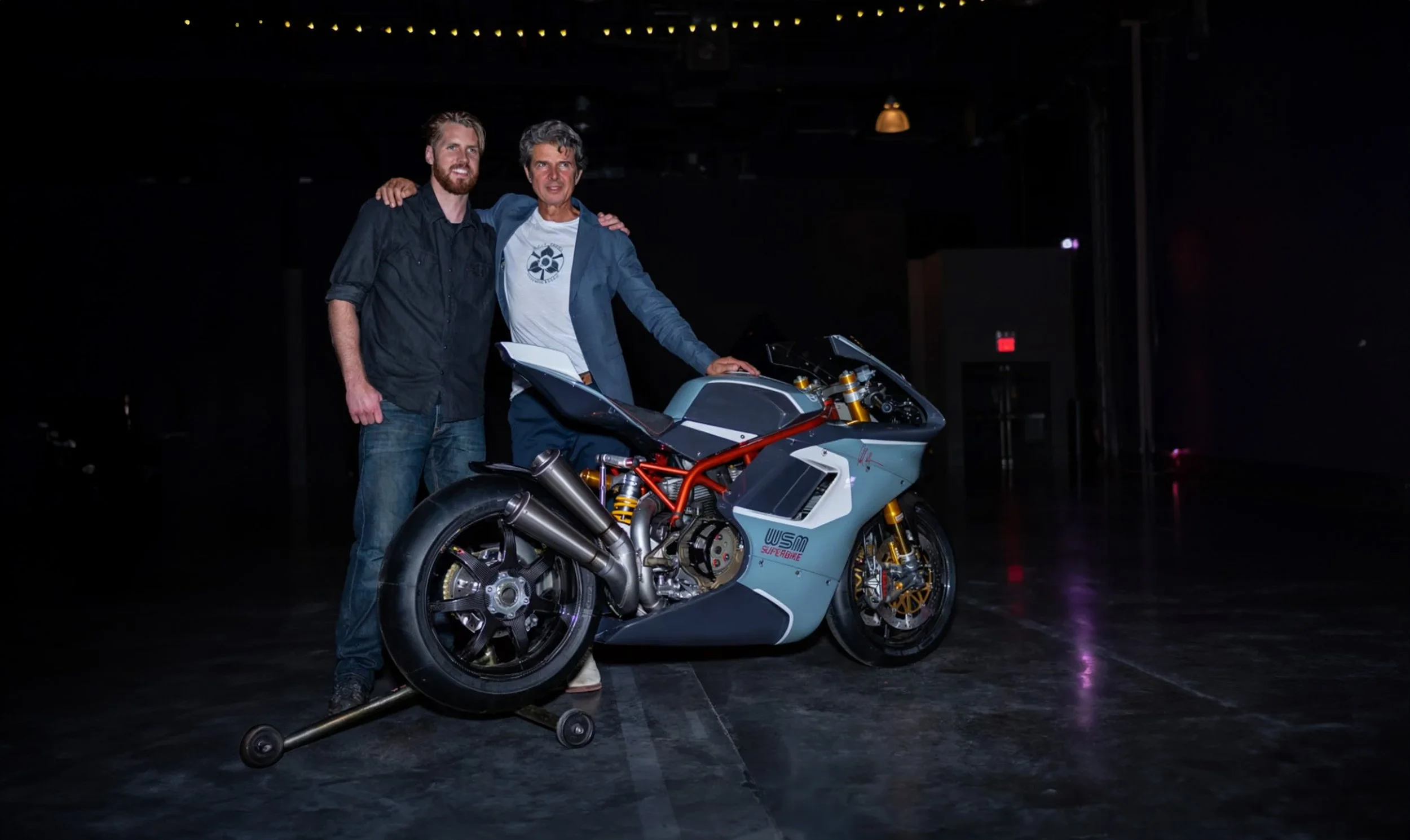 Two men standing next to a racing motorcycle on a dark stage, with one man resting his arm on the other's shoulder.