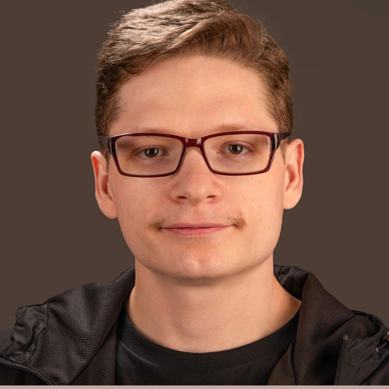 Close-up portrait of a young man with short brown hair, wearing glasses with dark red frames, and dressed in a black shirt with a black jacket, against a neutral background.