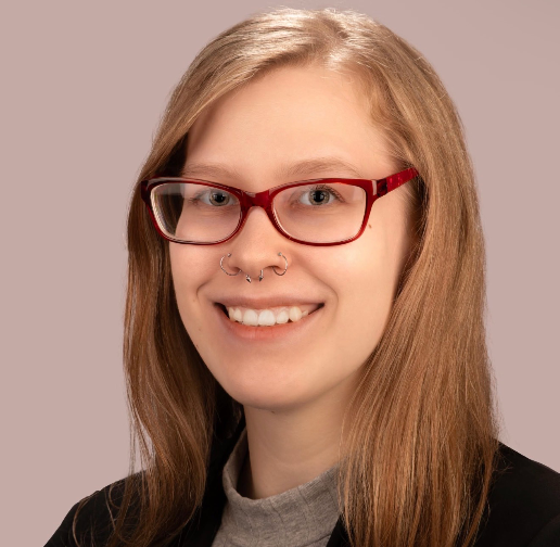 A woman with long red hair, red glasses, and a nose ring, smiling at the camera against a neutral background.