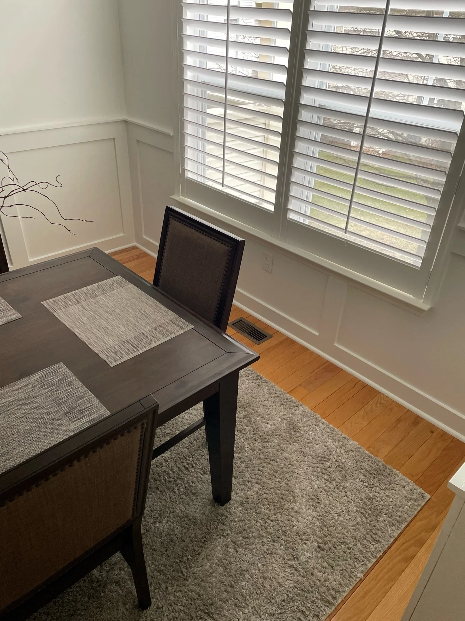 A dining room corner with a dark wooden table, beige upholstered chairs, window blinds, a gray area rug, and a view of a suburban yard outside.
