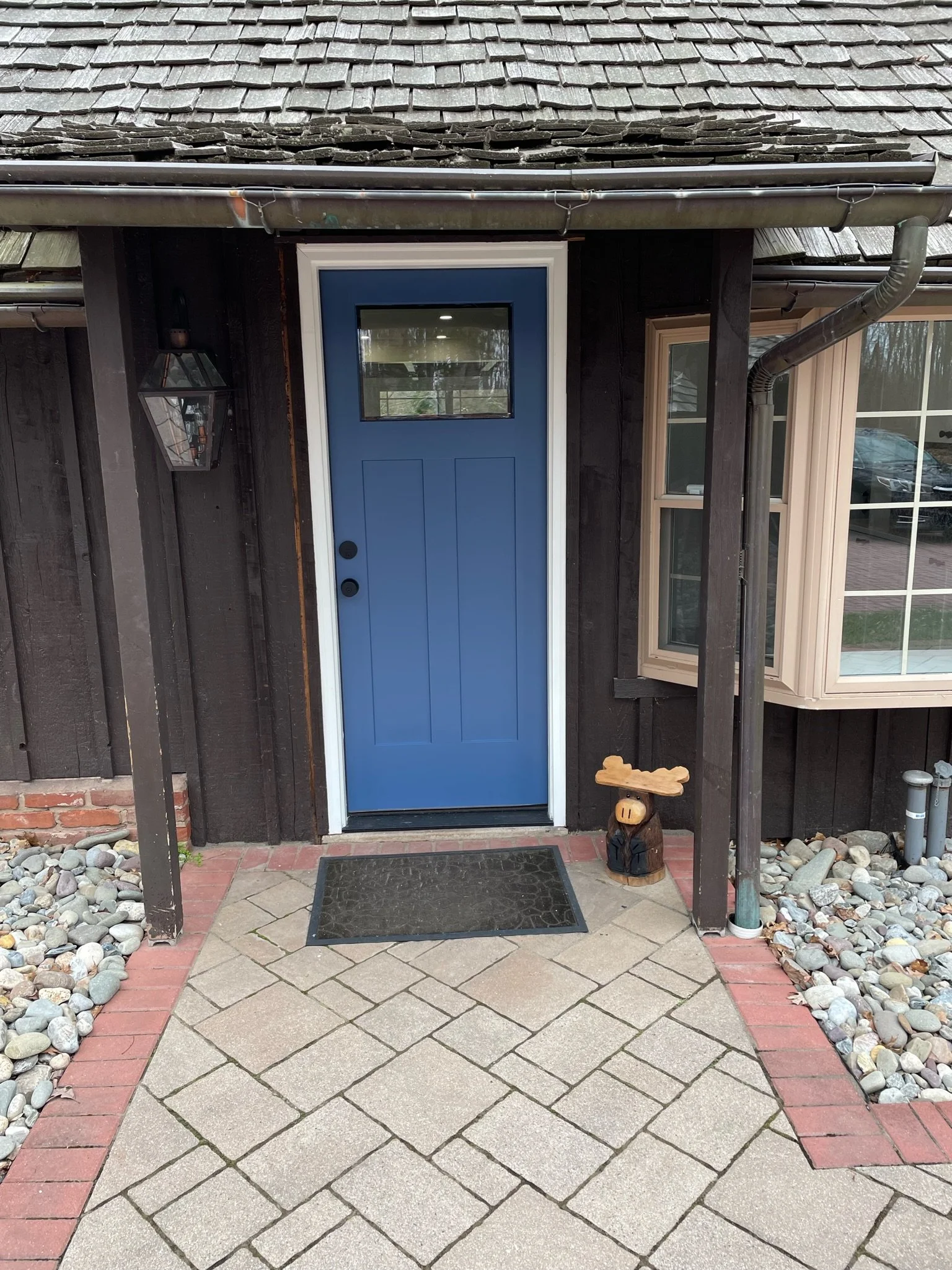 Front door of house with blue door, small window at top, black doorknobs, black mat, decorative pig figure on right, stone pathway, rock landscape on sides, dark wood siding, window on right, lantern light on left, gutter system.