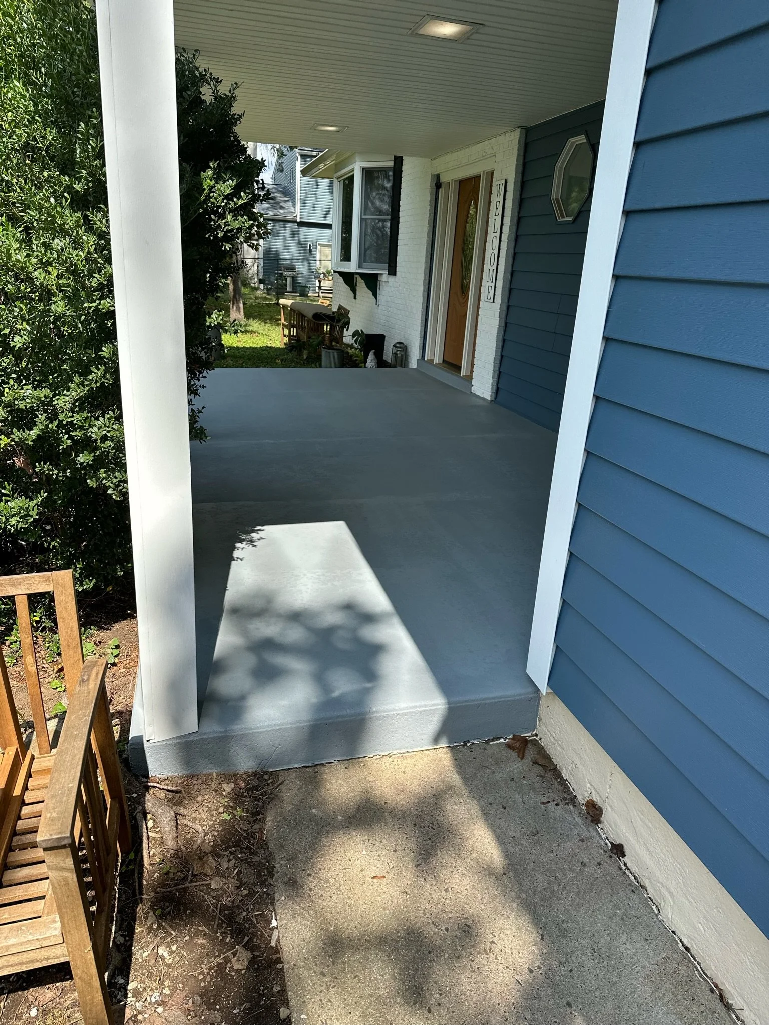 Front porch of a house with a blue exterior wall, a concrete pathway, and a welcome sign near the front door. There are trees, bushes, and outdoor furniture visible in the background.