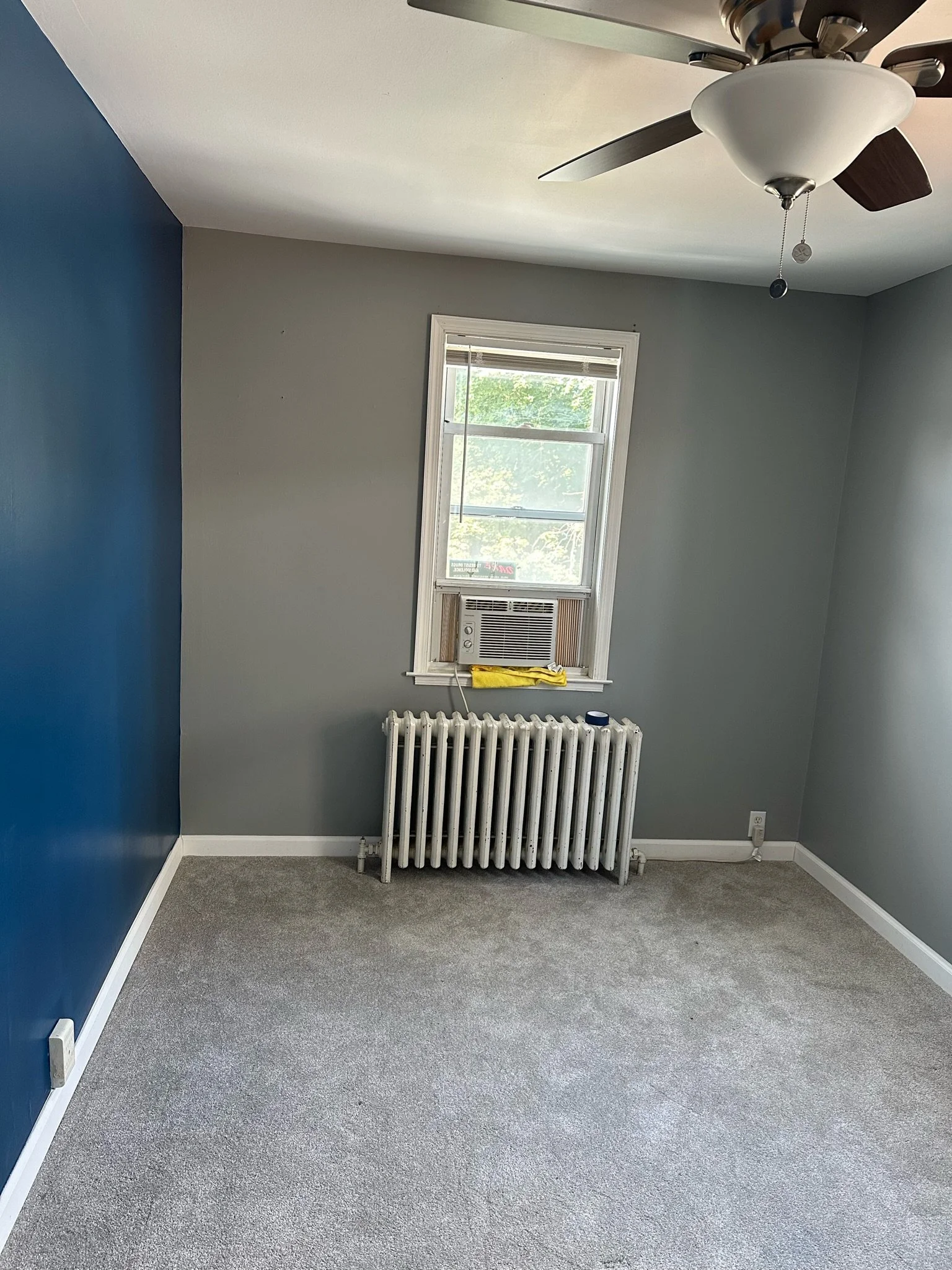Empty room with gray walls, a dark blue accent wall, gray carpet, a window with an air conditioning unit, a white radiator beneath the window, and a ceiling fan with light.