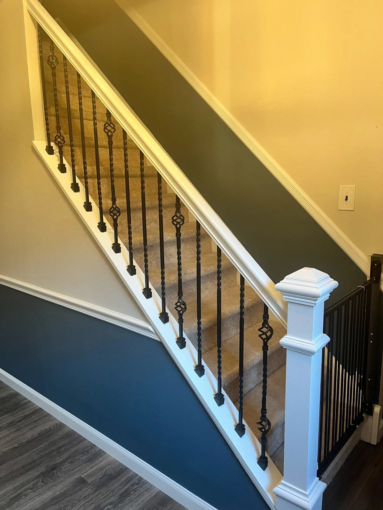 Interior staircase with beige carpeting, black wrought iron balusters, white railing and trim, and dark blue wall underneath.
