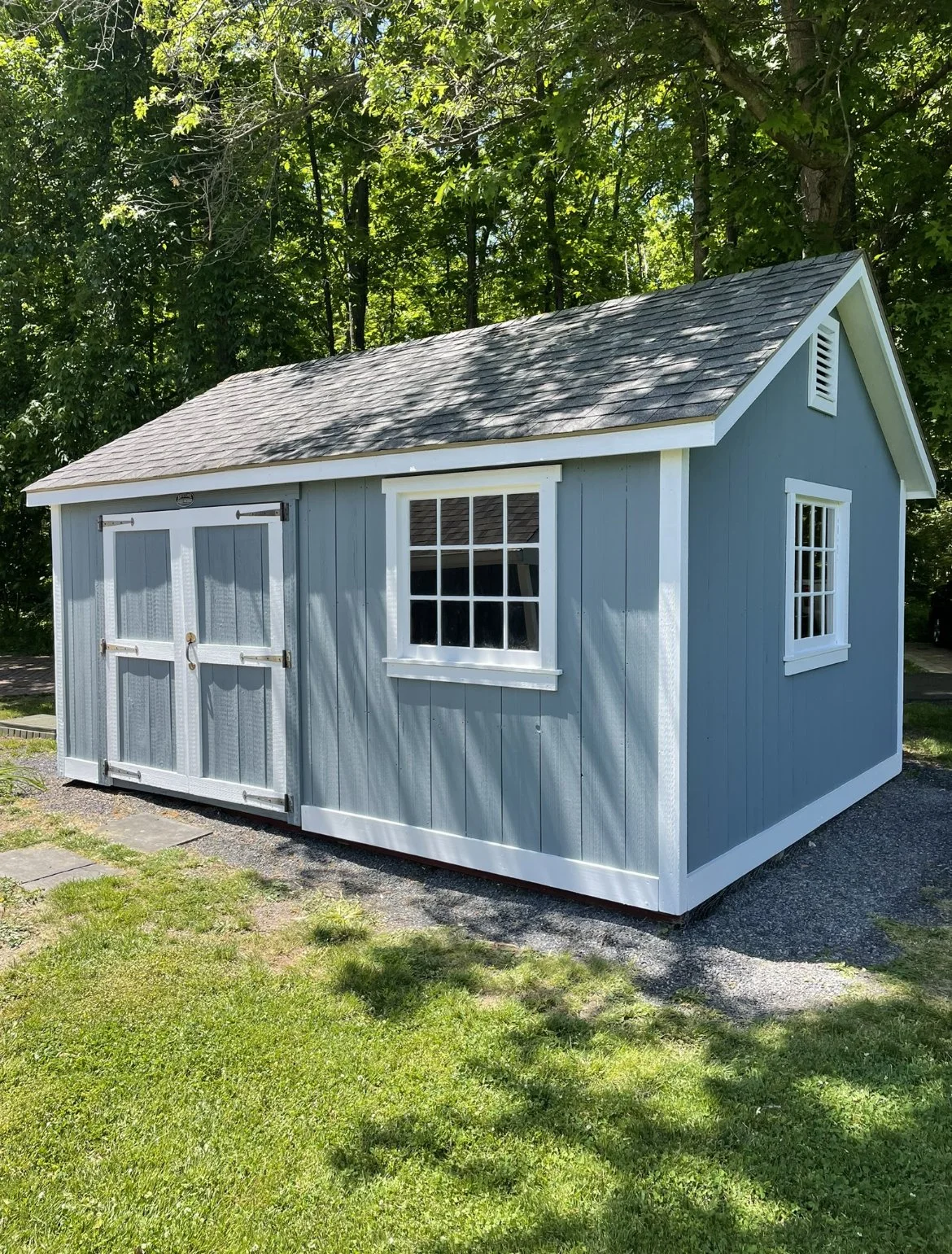 A small blue garden shed with white trim and a shingled roof, situated on a grassy lawn with trees in the background.