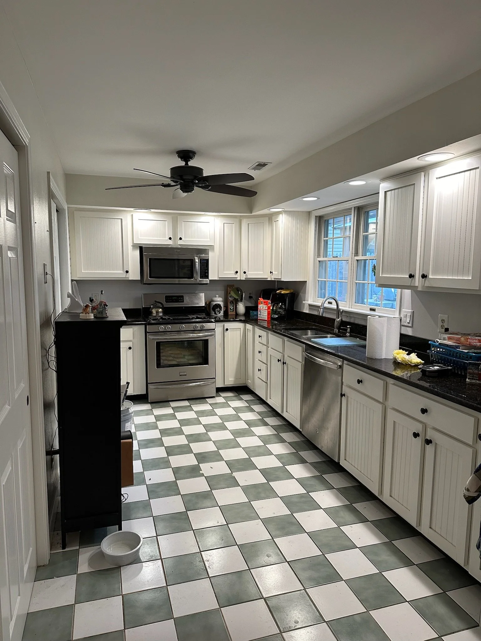 Kitchen with white cabinets, black countertop, stainless steel appliances, checkered green and white floor, and a ceiling fan.