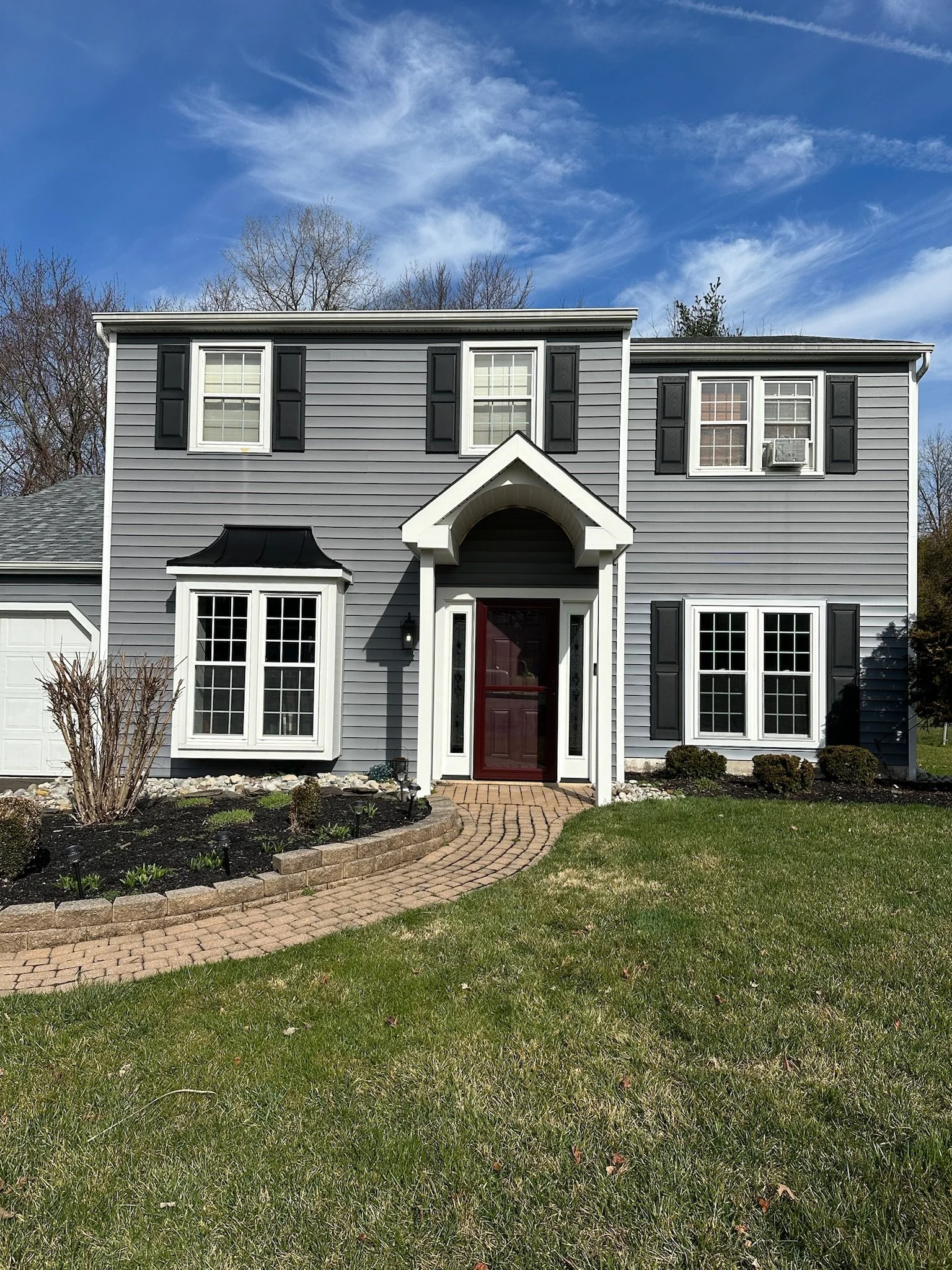 Front view of a two-story house with gray siding, black shutters, white trim, a brick walkway, a small front yard with grass and bushes, and a blue sky with clouds.