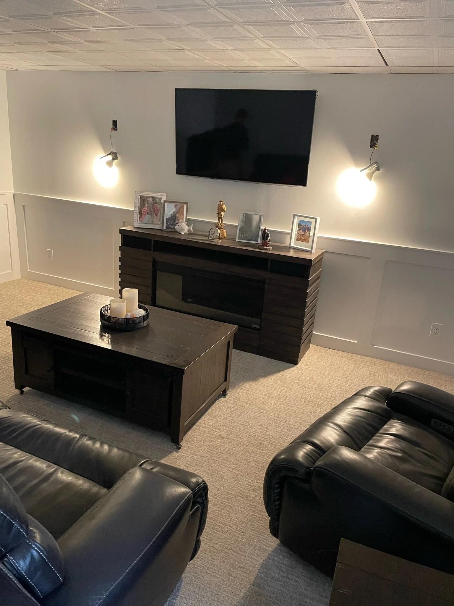 A cozy living room with two black leather sofas, a wooden coffee table with candles, a wall-mounted TV, and a dark wood entertainment center decorated with framed photos, a small statue, and a clock. Wall sconces provide lighting.
