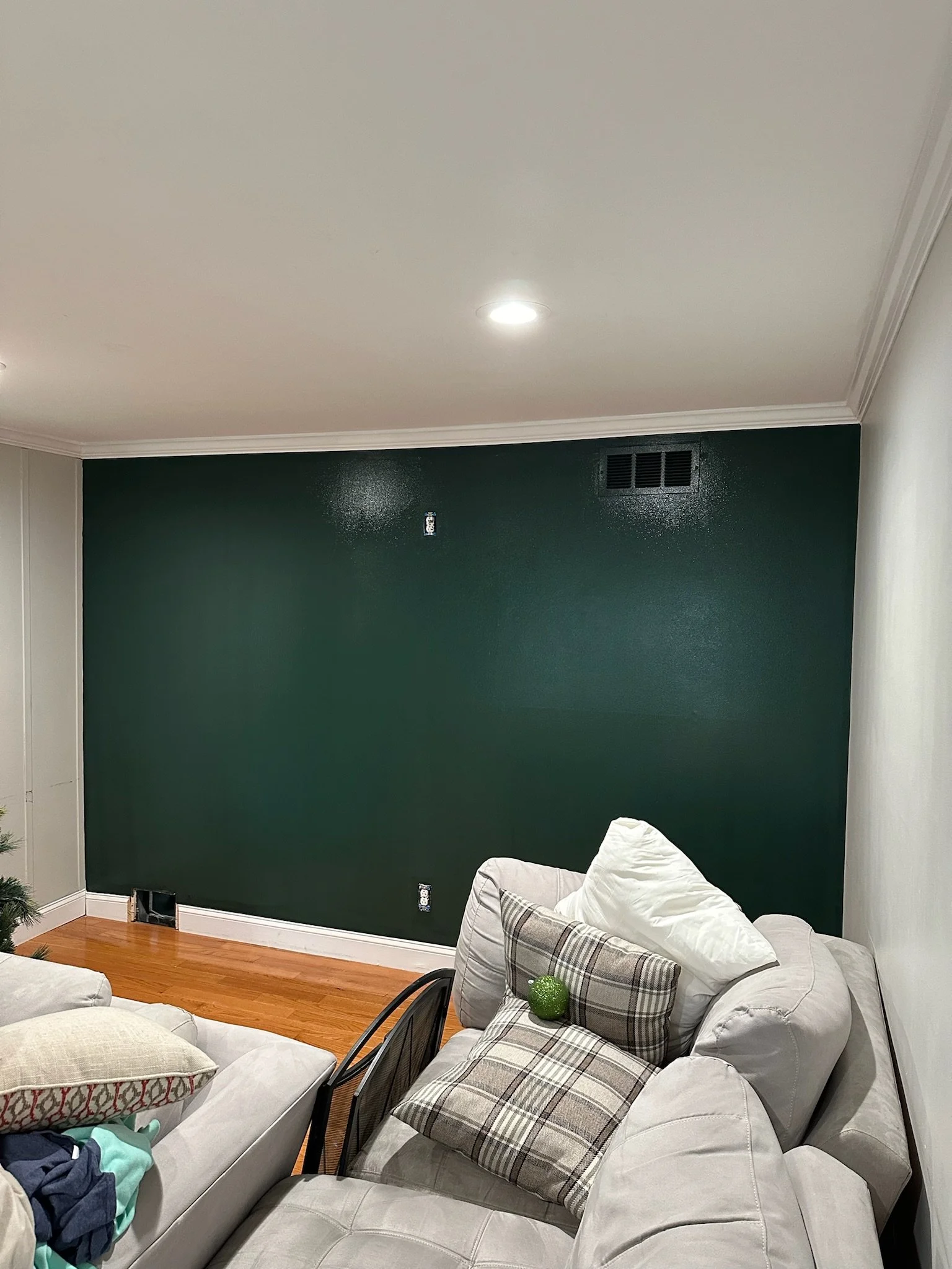 Living room with a green accent wall, white ceiling, hardwood floor, beige sectional sofa with various pillows, and some clothing items on the sofa.