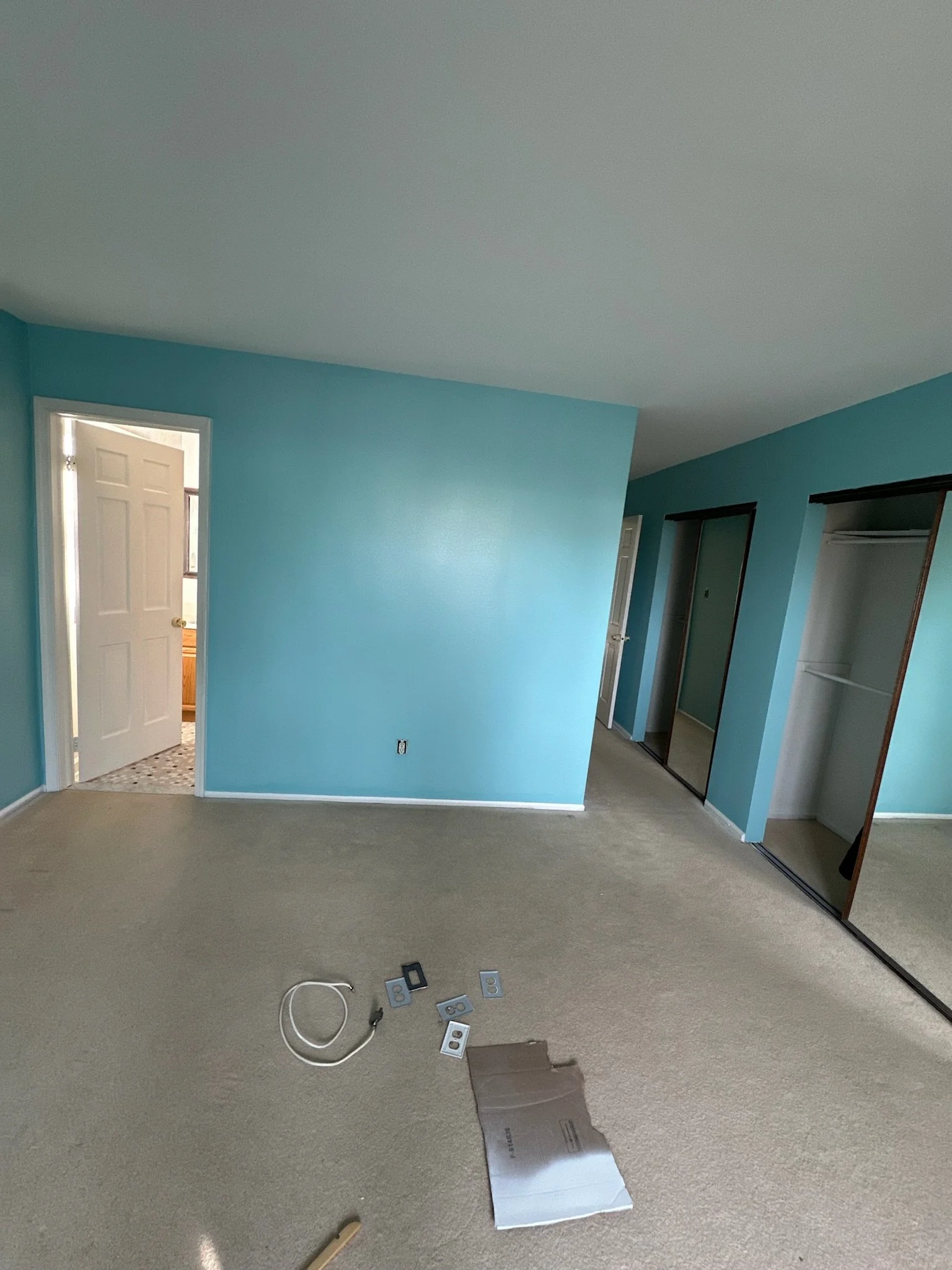 Empty bedroom with blue walls, an open closet with sliding mirror doors, a door leading to another bathroom, and some electrical fixtures and tools on the beige carpeted floor.
