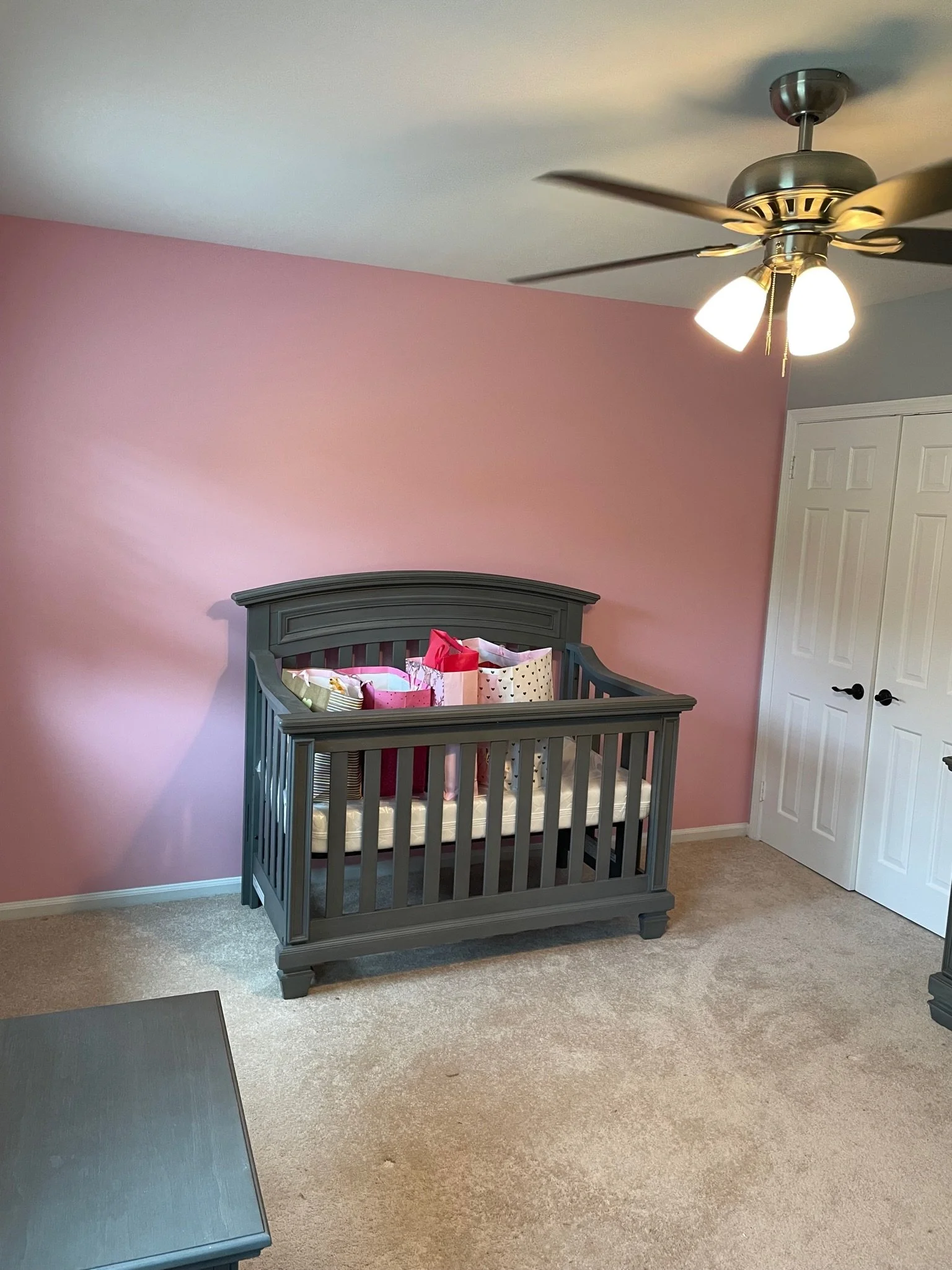 A gray crib with pink, white, and patterned bedding set against a pink accent wall in a nursery. There is a ceiling fan with three lights above and a white closet with double doors on the right. The floor is beige carpet.
