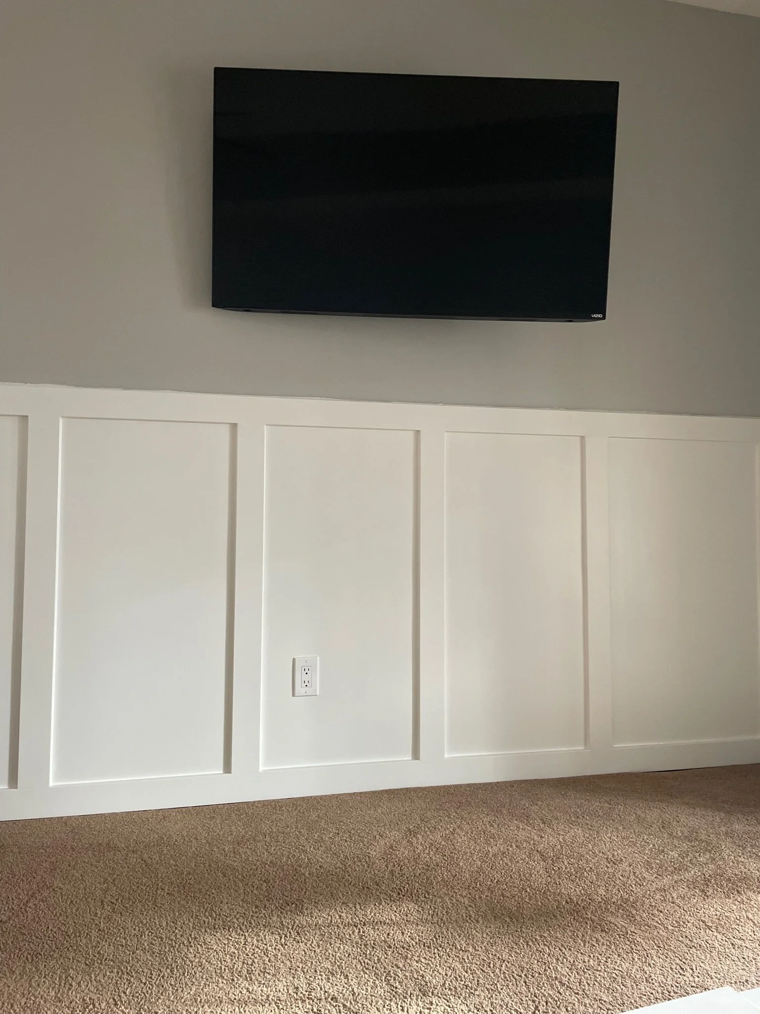 Empty room with a mounted flat-screen television, white wall paneling, electrical outlet, and beige carpet.