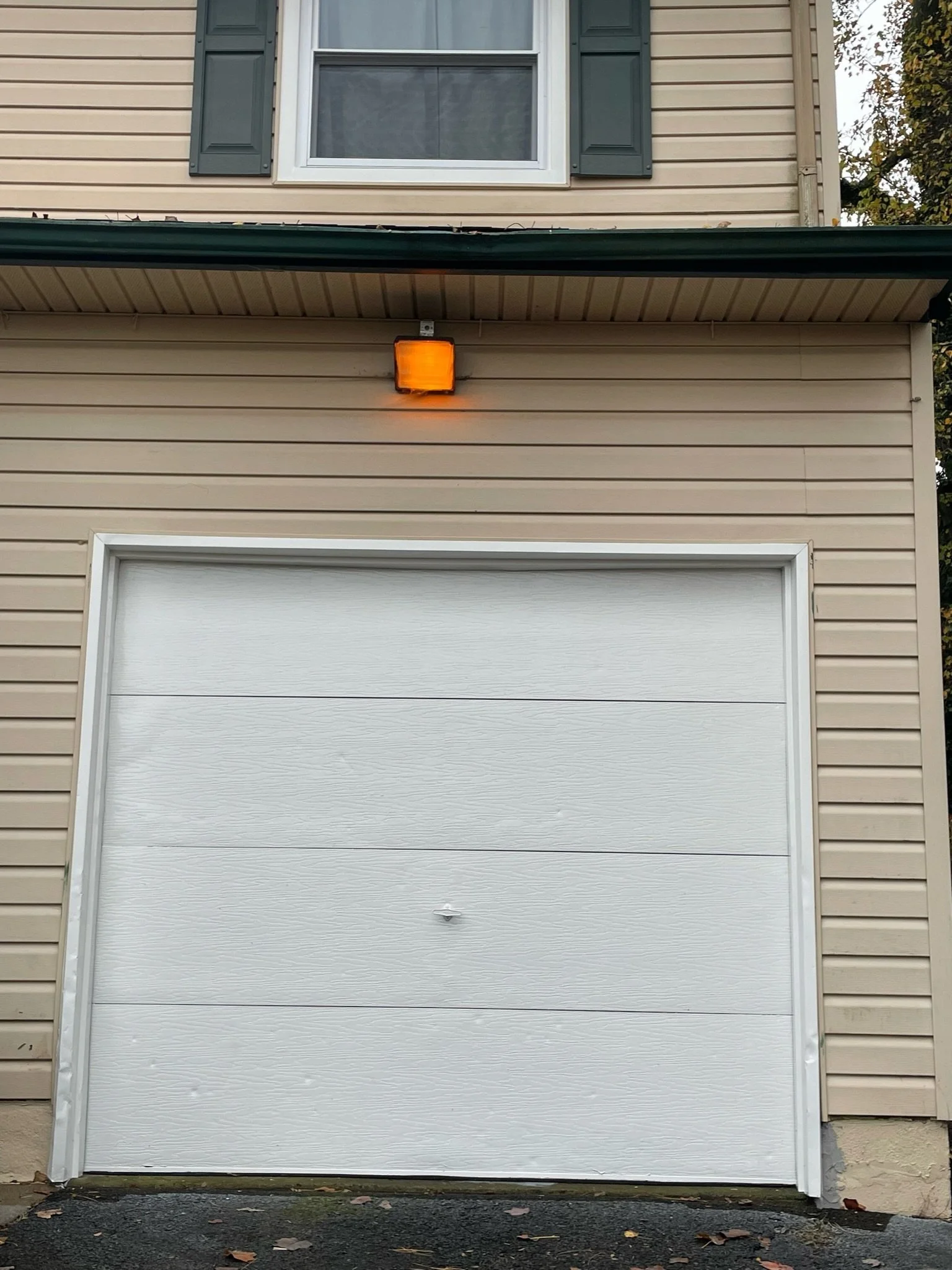 Residential garage door with beige siding, white door, a small window above with dark green shutters, and an orange exterior light mounted above the garage.