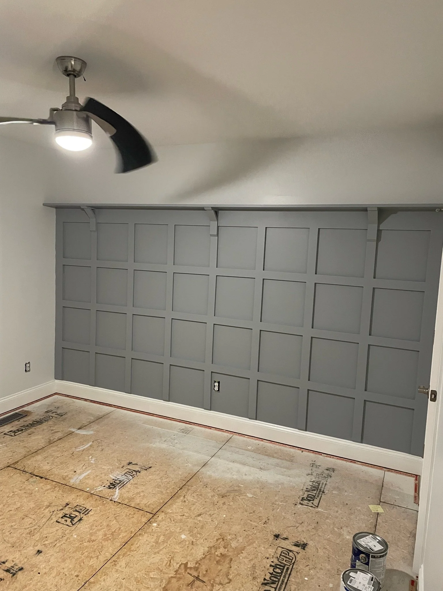 Unfinished basement room with gray paneled wall, drywall, and a ceiling fan with a light fixture. Partial floor covered with plywood sheets for flooring and paint cans on the floor.