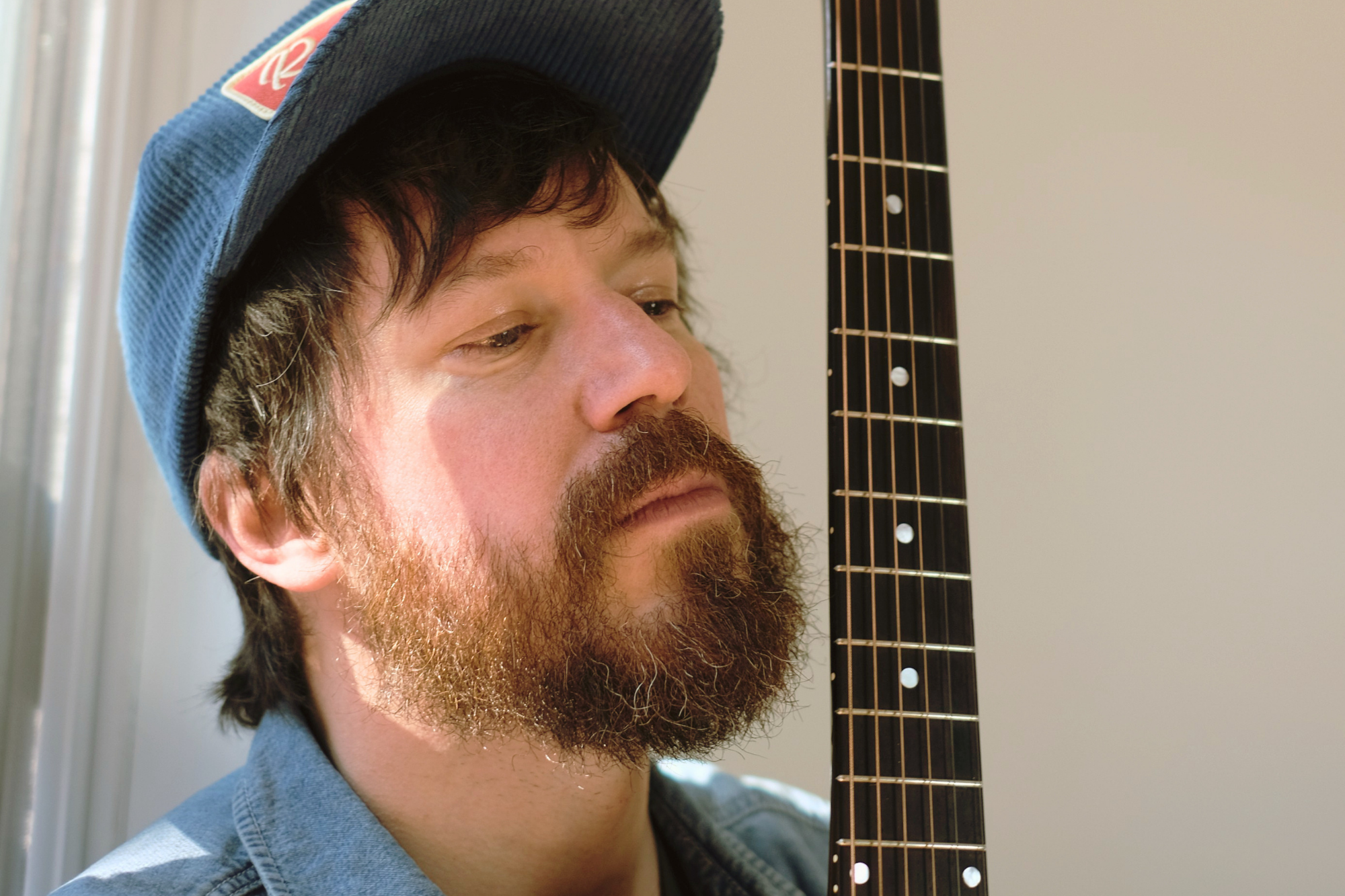 A man with a beard and mustache wearing a blue hat and denim shirt, holding a guitar close to his face.