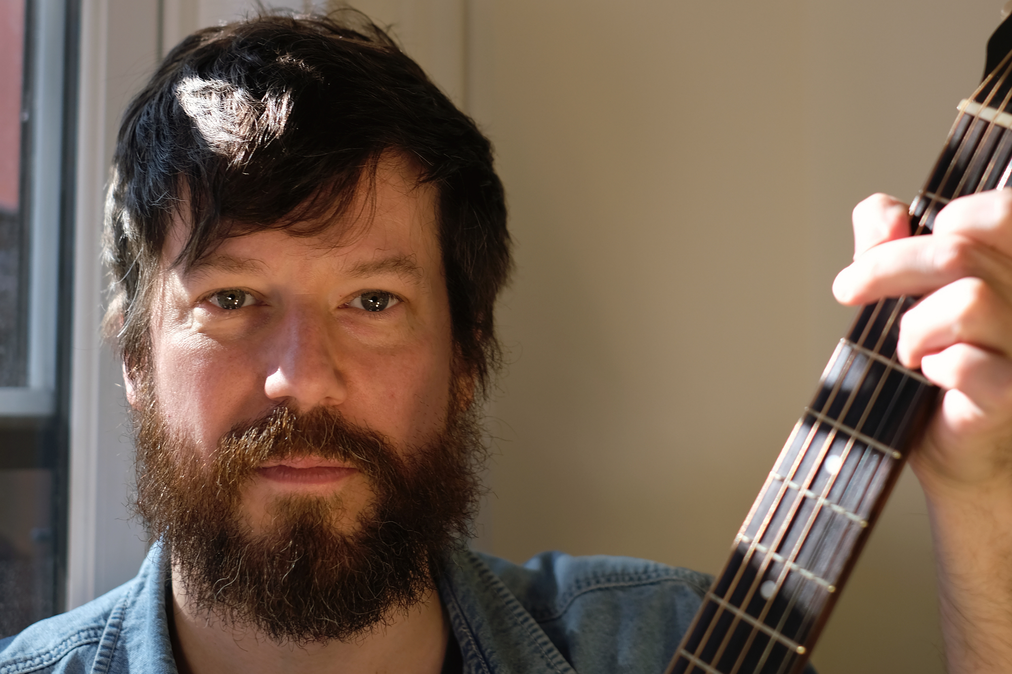 A man with dark hair and a beard holding a guitar near his face, looking at the camera, standing by a window.