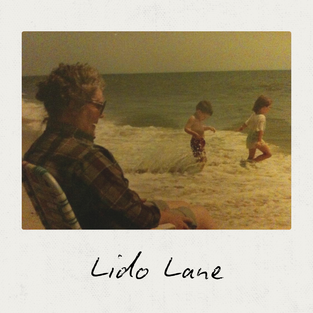 A person sitting on a beach chair near the ocean watching two children playing in the shallow water at the shoreline, with waves rolling in. The image has a nostalgic, vintage feel. The caption reads "Lido Lane."
