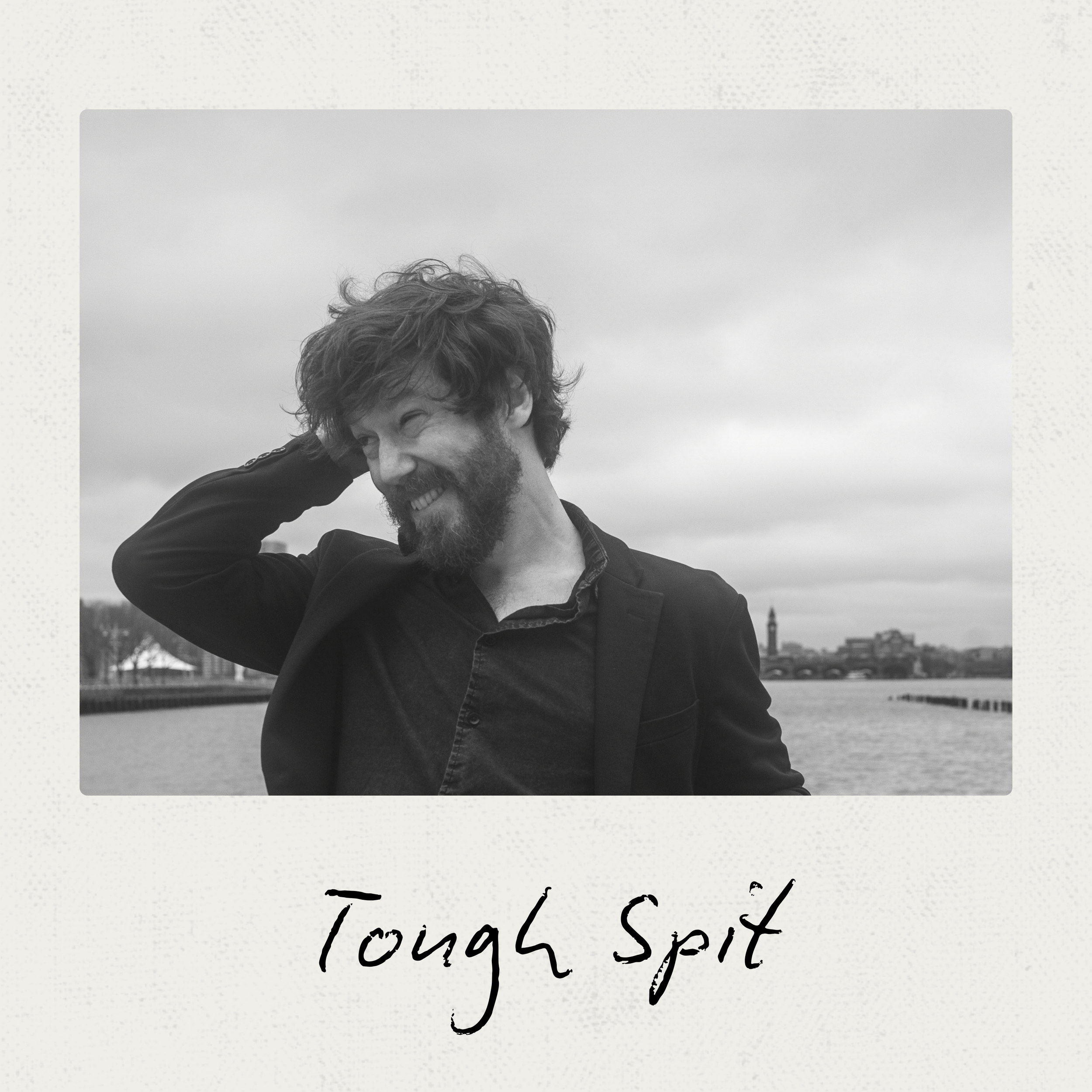 A black and white photo of John smiling and scratching his head outdoors near a city waterfront.