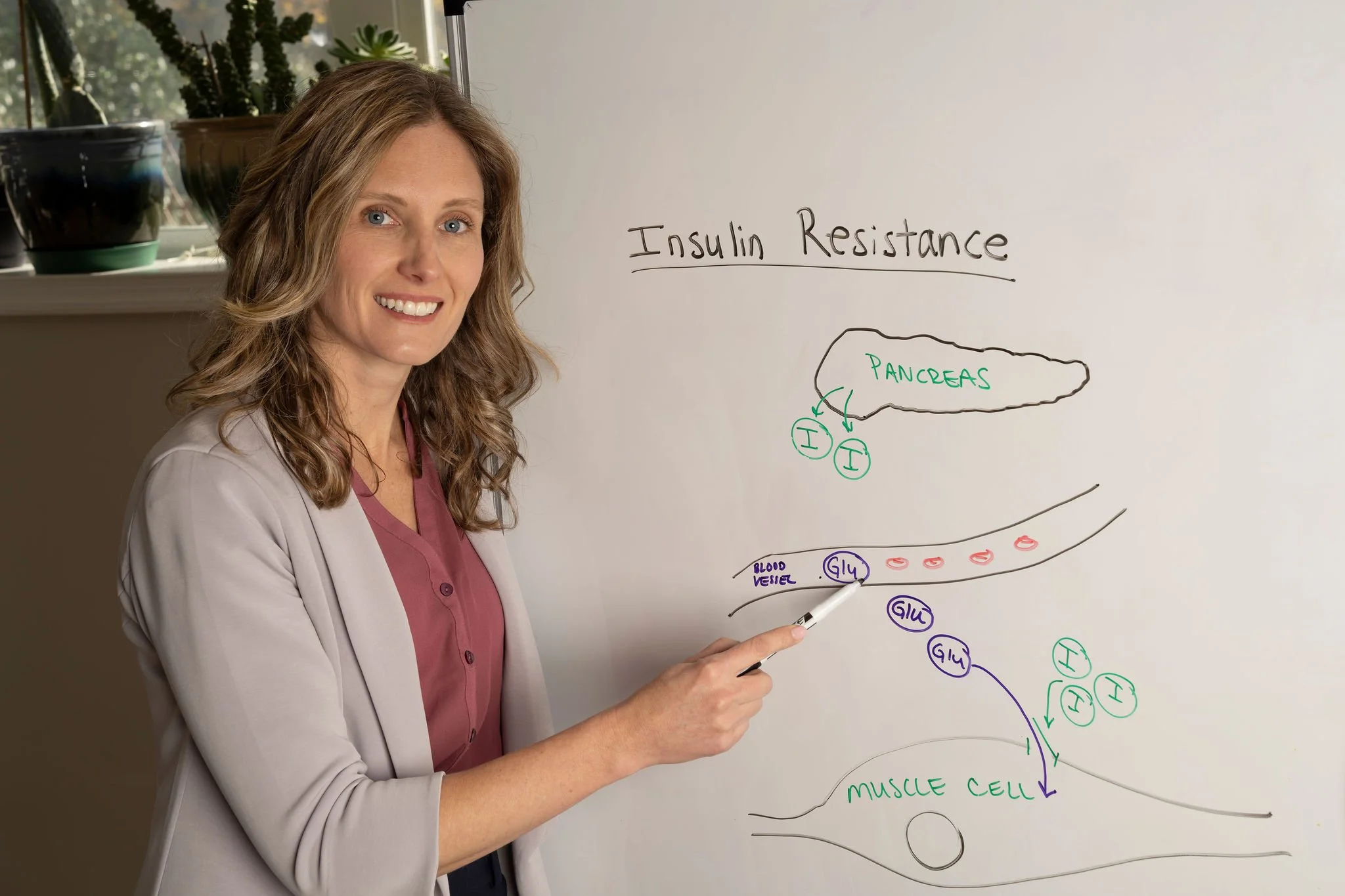 A woman in a blazer and maroon shirt standing next to a whiteboard with diagrams and notes about insulin resistance, pointing at the board with a marker.