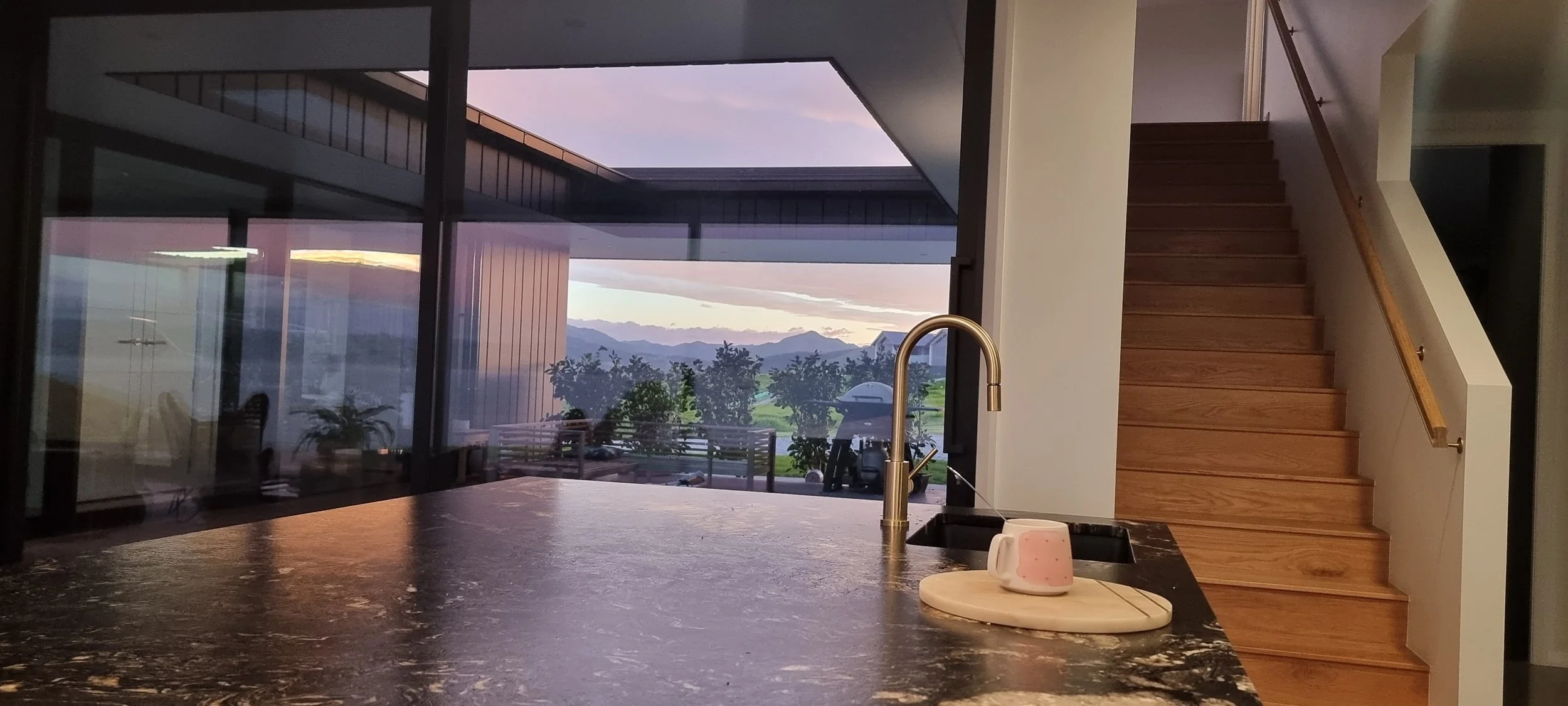 Modern kitchen interior with a marble countertop, a gold faucet, and a view of mountains and trees outside through large glass windows. Wooden staircase on the right side.