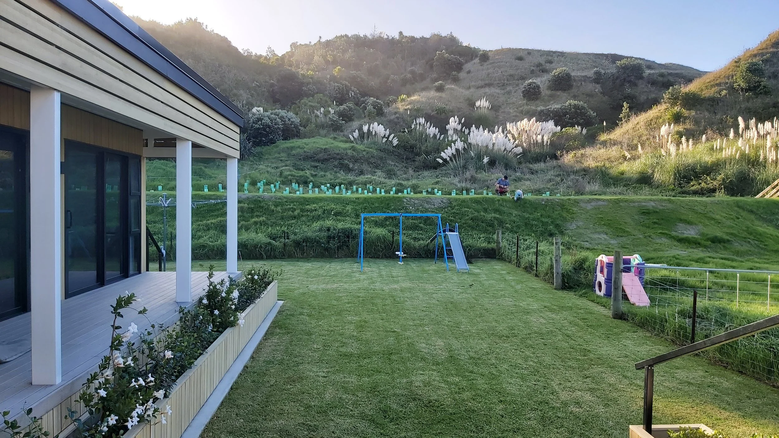 Backyard with green grass, a playset including a swing set and slide, a fenced area, and a hillside with tall grass and bushes in the background. Part of a modern house with large glass doors is on the left.