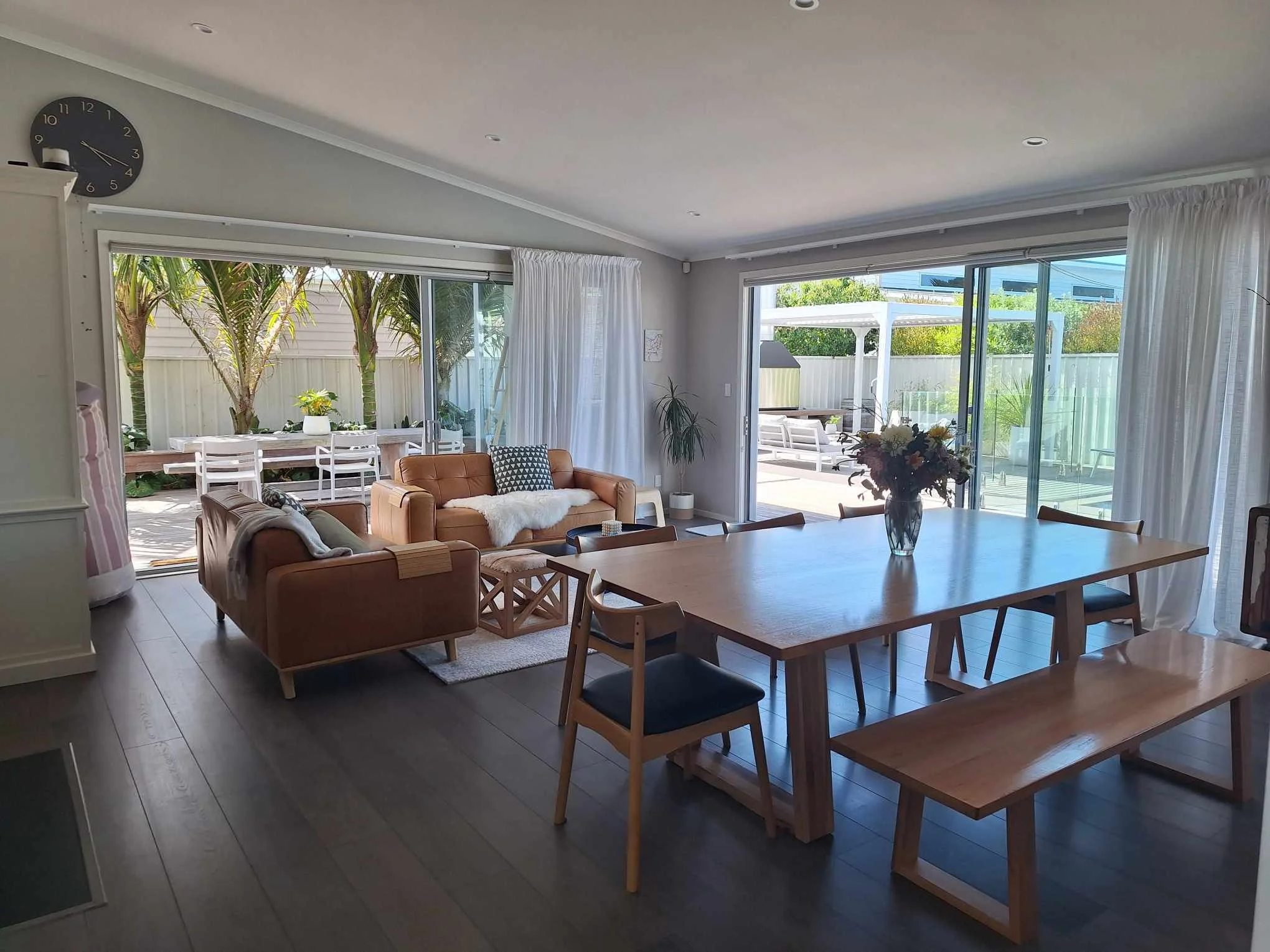 A bright living room with large sliding glass doors leading to an outdoor patio with seating. Inside, there are tan couches, a wooden dining table with chairs, and a vase with flowers. The room has white curtains, a wall clock, and wooden flooring.