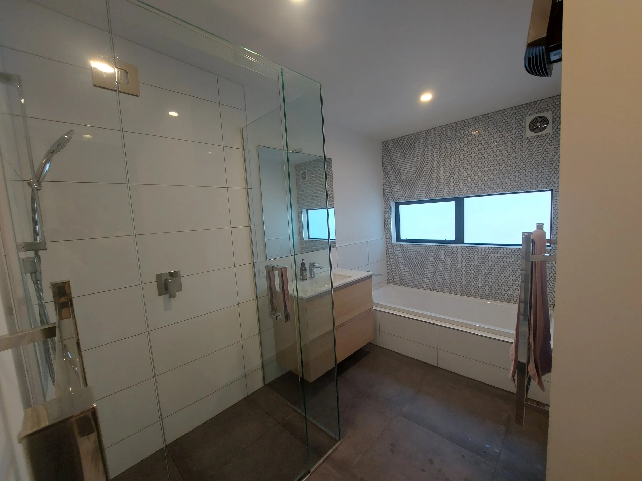 Modern bathroom with glass shower enclosure, white tiled walls, and a vanity with a sink. There are two windows and ceiling lights, with a towel hanging on a rail.